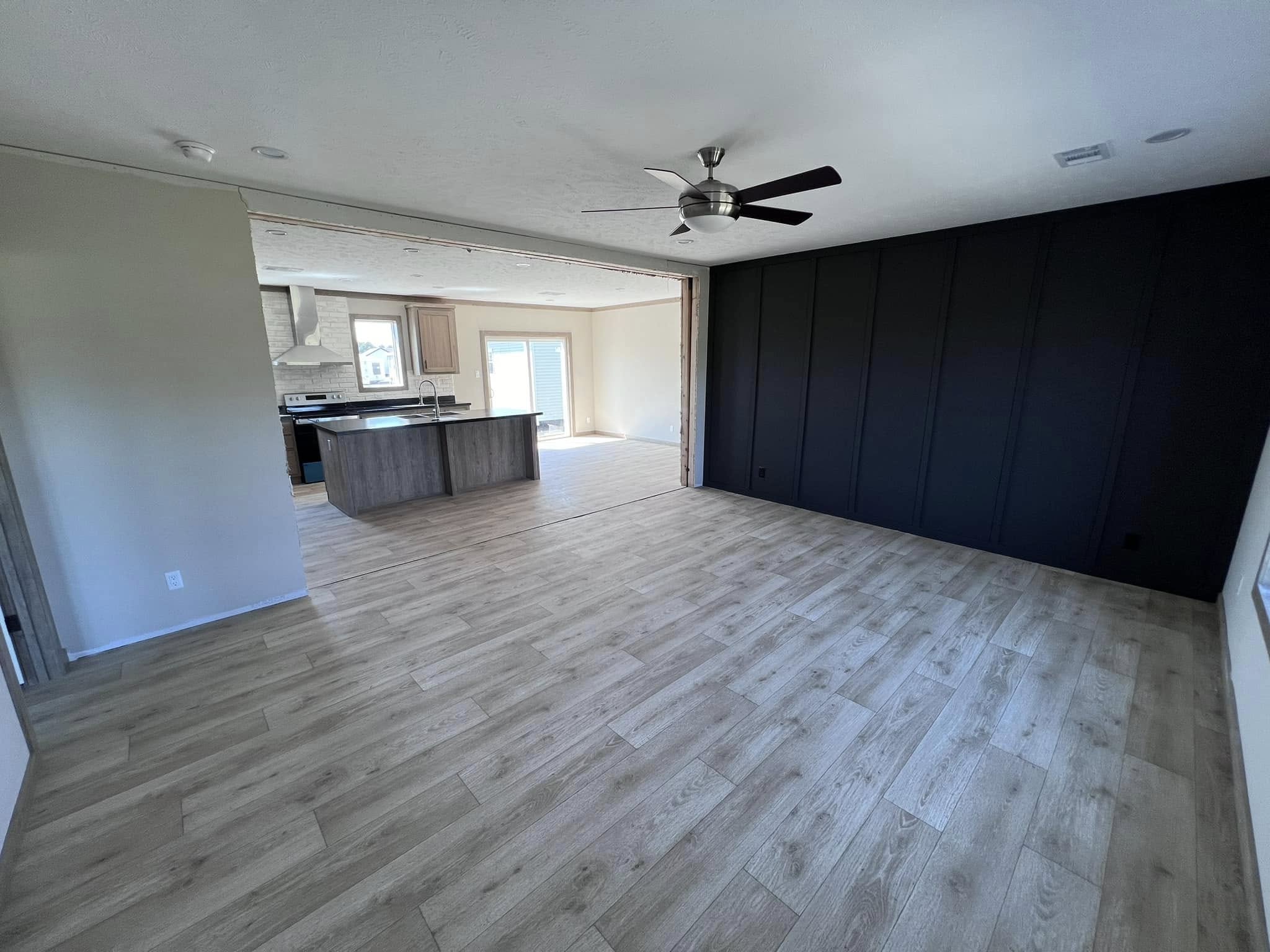 Spacious open-plan room with light wood flooring, a black accent wall, and a ceiling fan. Kitchen area in the background with an island and patio doors.
