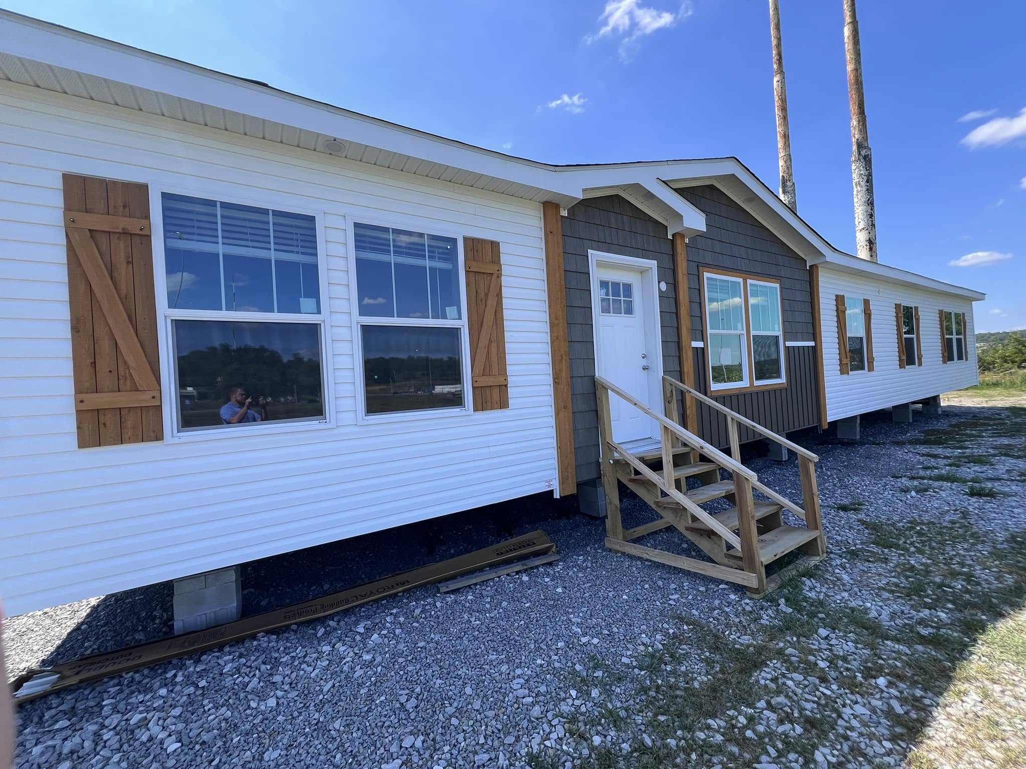 A modular home with white siding and brown accents sits on a gravel lot under a clear blue sky. Wooden steps lead to a white door, exuding a simple, rustic charm.