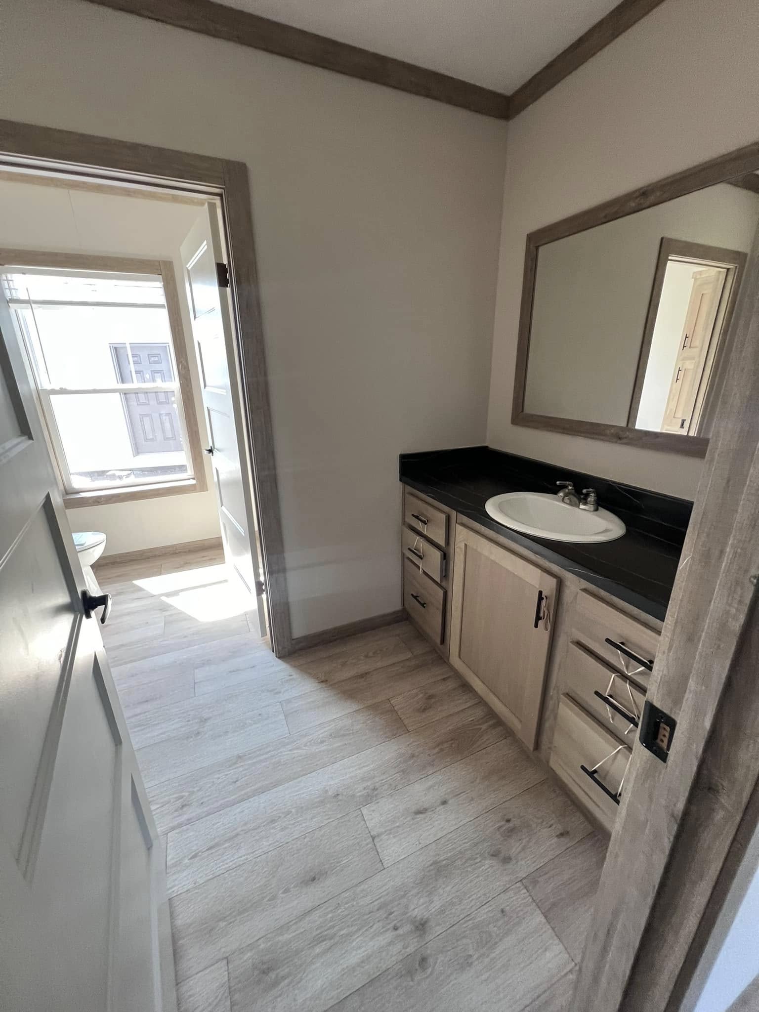A wooden-floored bathroom with a light wood vanity, black countertop, and white sink. A large mirror hangs above. Natural light streams from a window.