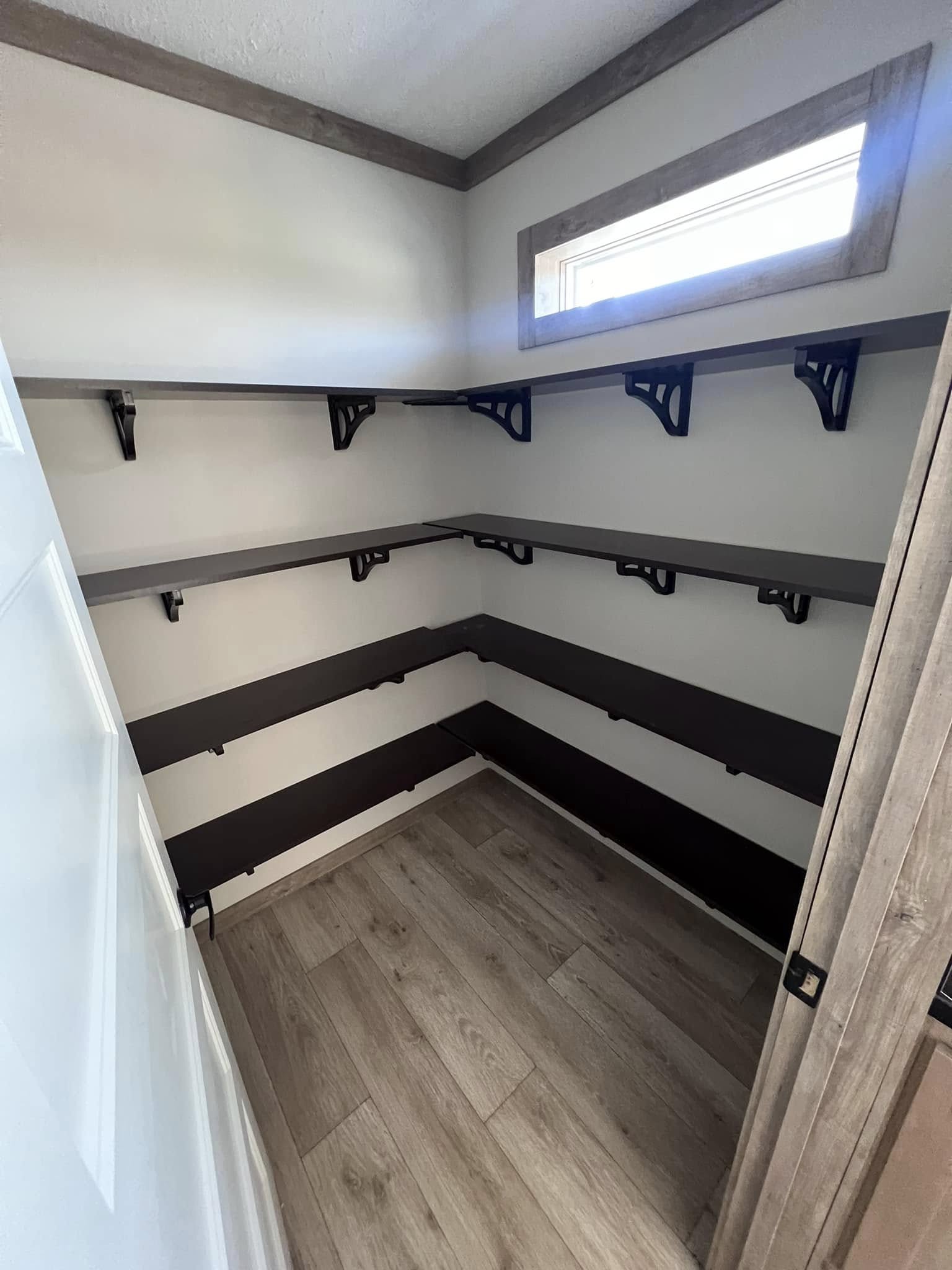 Small, empty pantry with wooden shelves and light wood flooring. The space has a simple, tidy appearance with ample shelving and natural light.