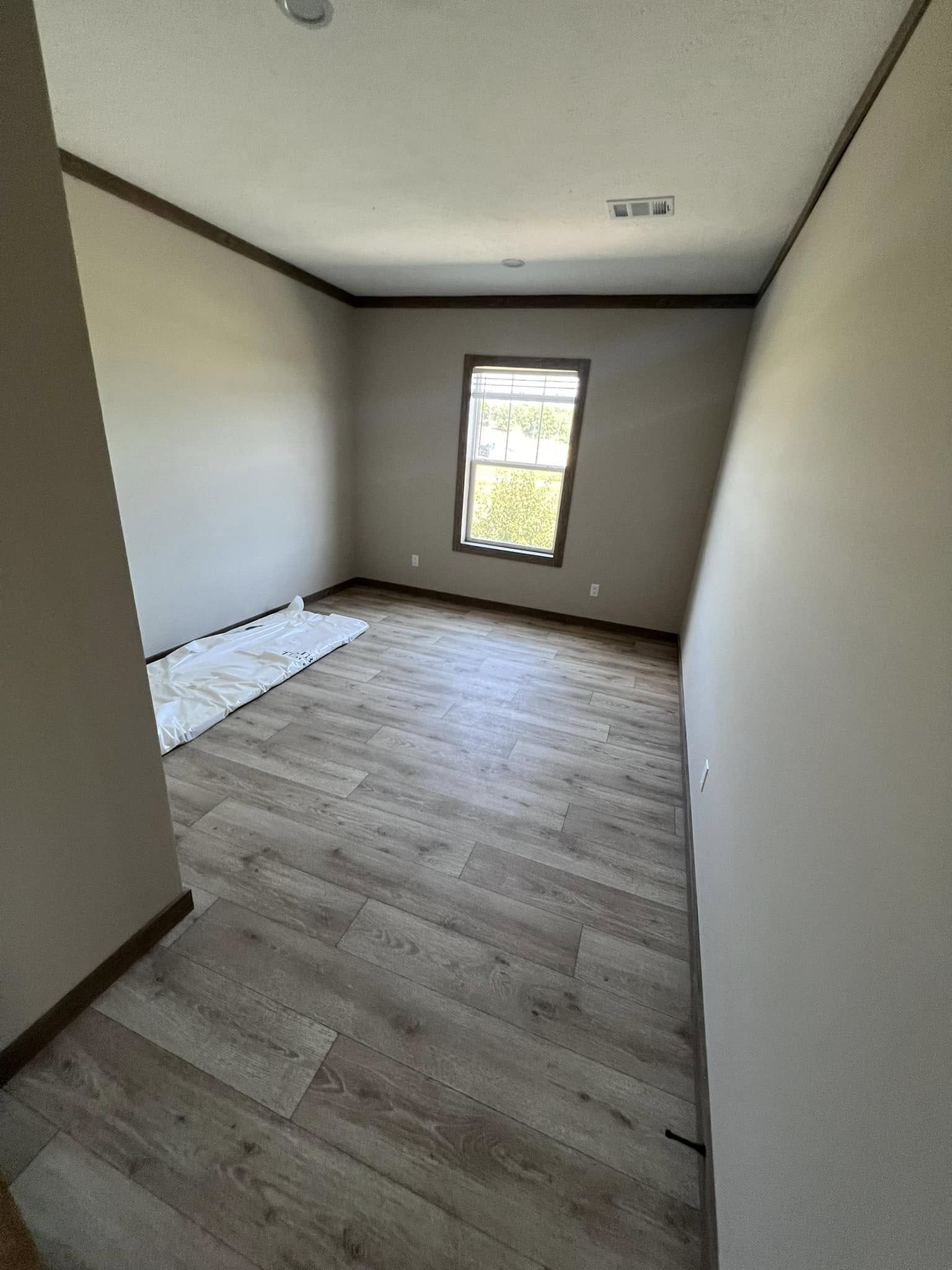 Empty room with light wood flooring, beige walls, and a single window letting in natural light. A folded white sheet lies on the floor, creating a calm, minimalistic atmosphere.