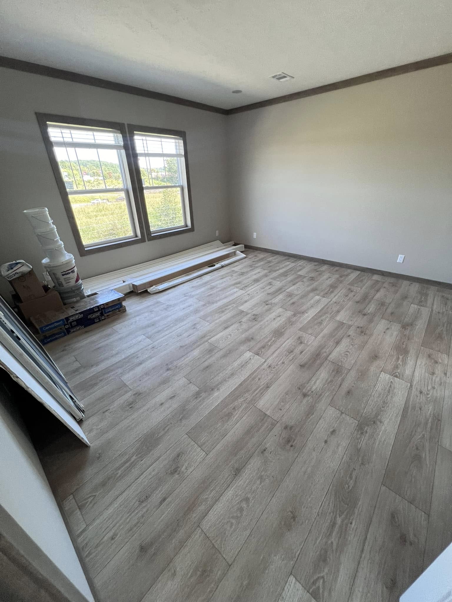 An empty room with unfinished laminate wood flooring, beige walls, and a large window. Construction materials are stacked near the window.