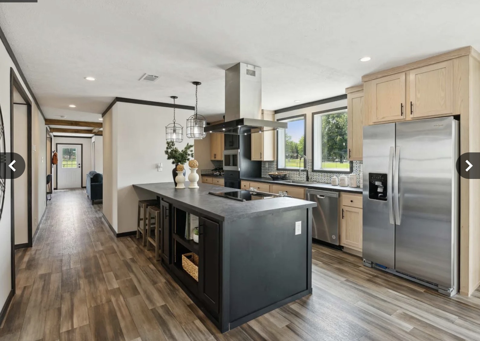 Modern kitchen with wood flooring, featuring a central black island with stools, pendant lights, light wood cabinets, stainless steel appliances, and large windows for natural light.
