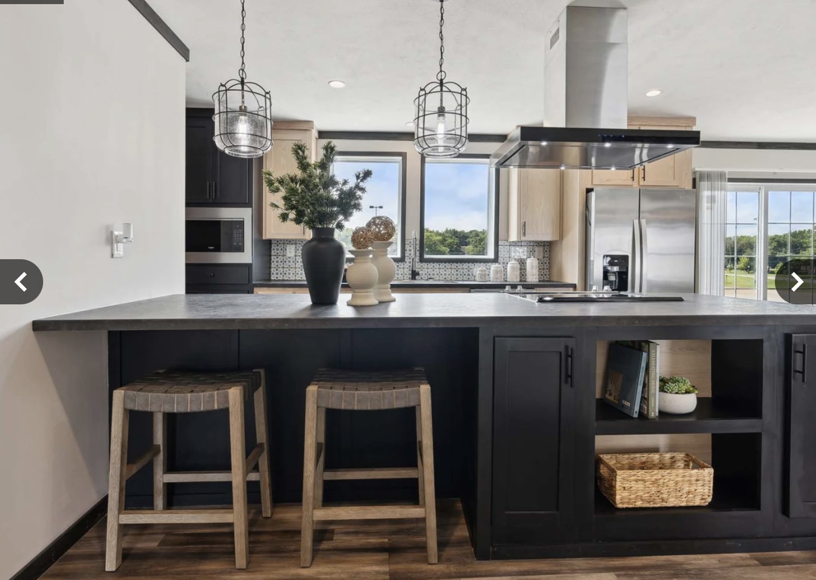 Modern kitchen with a large island featuring two wooden stools, pendant lights, and decorative vases. The space is bright and airy with large windows.