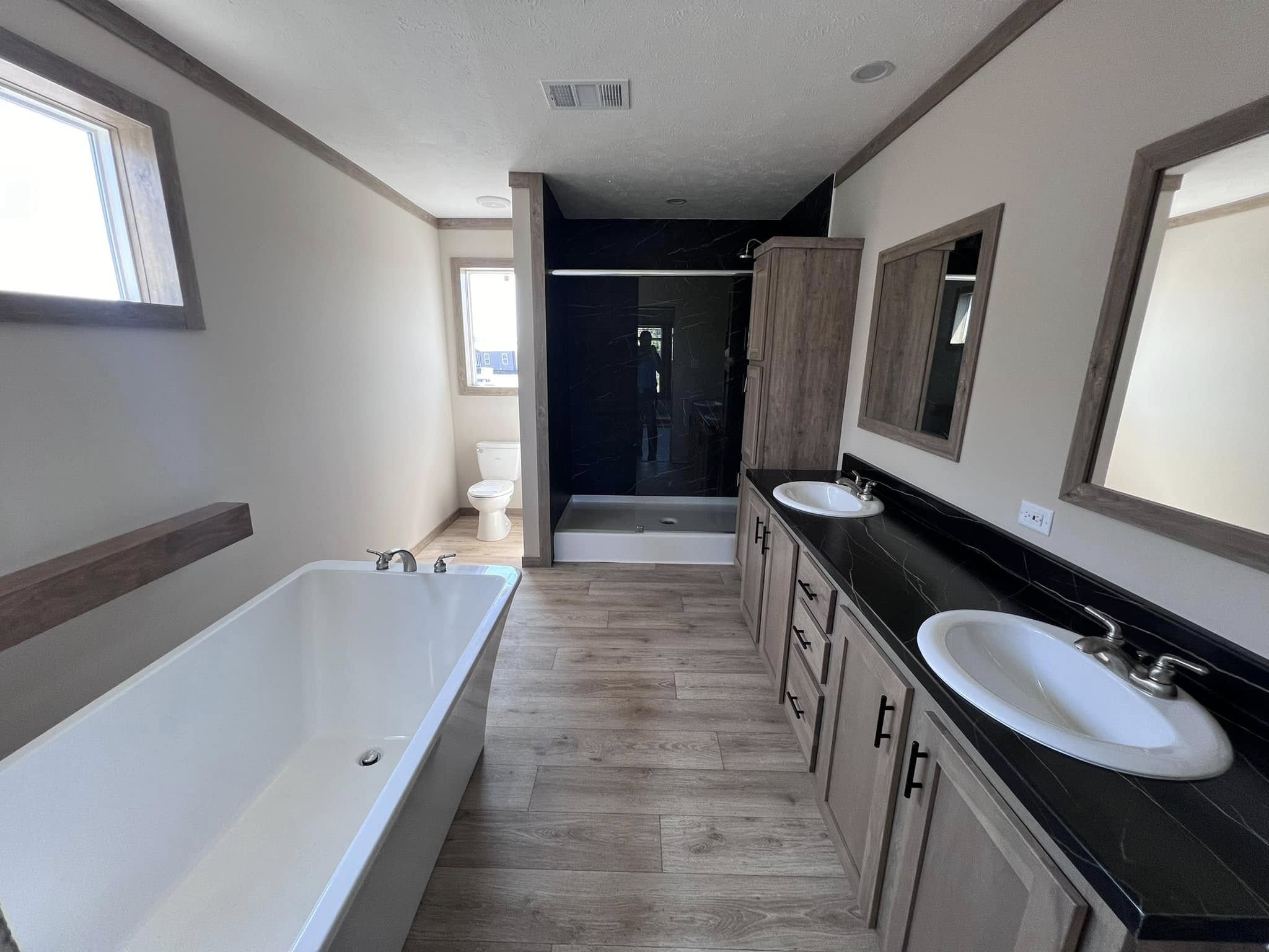 Spacious bathroom with light wood flooring, a large bathtub, twin sinks on a black countertop, and a glass shower. Bright and modern tone.