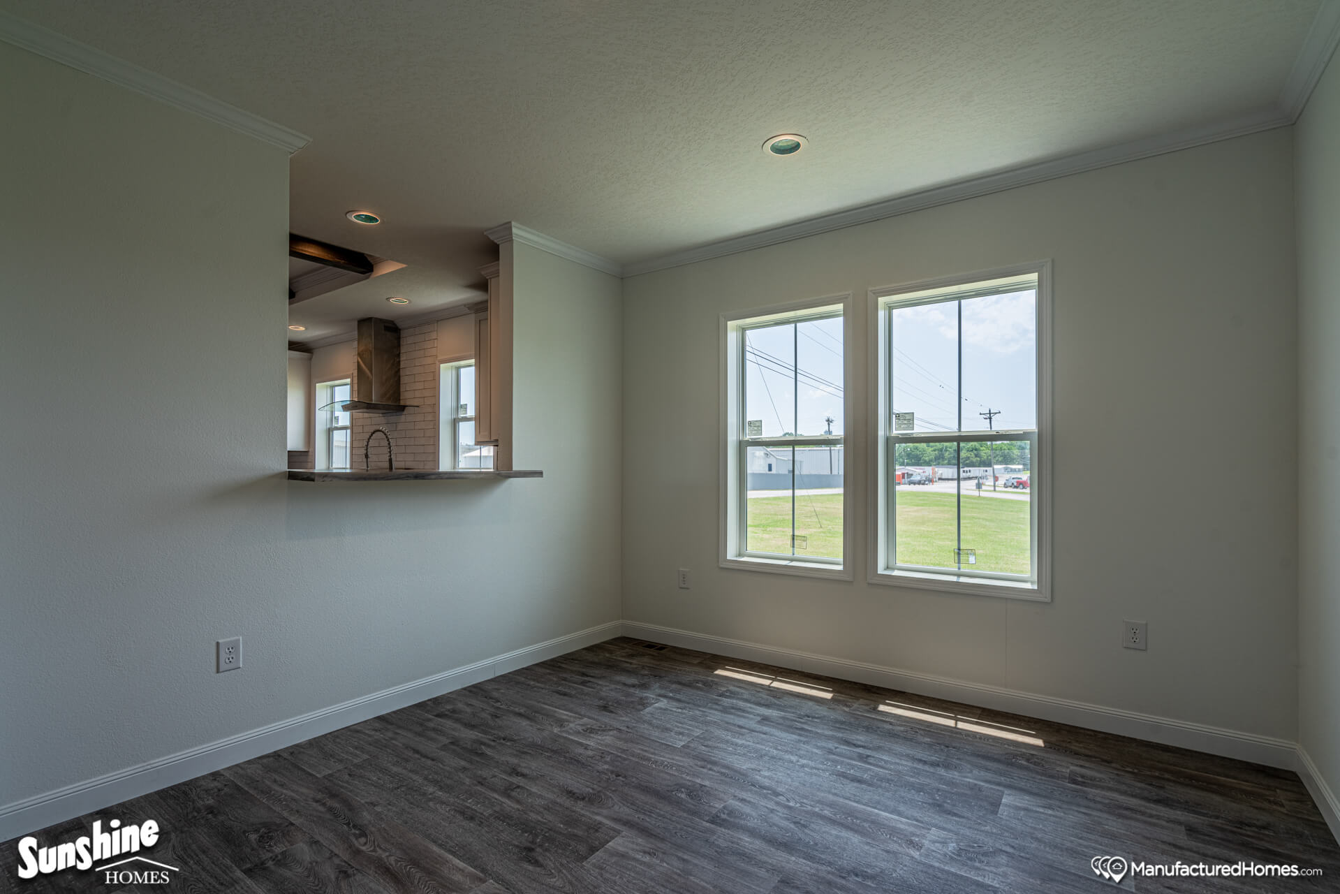 Bright room with dark wood laminate flooring, white walls, and two large windows offering an outside view. A kitchen pass-through is on the left.