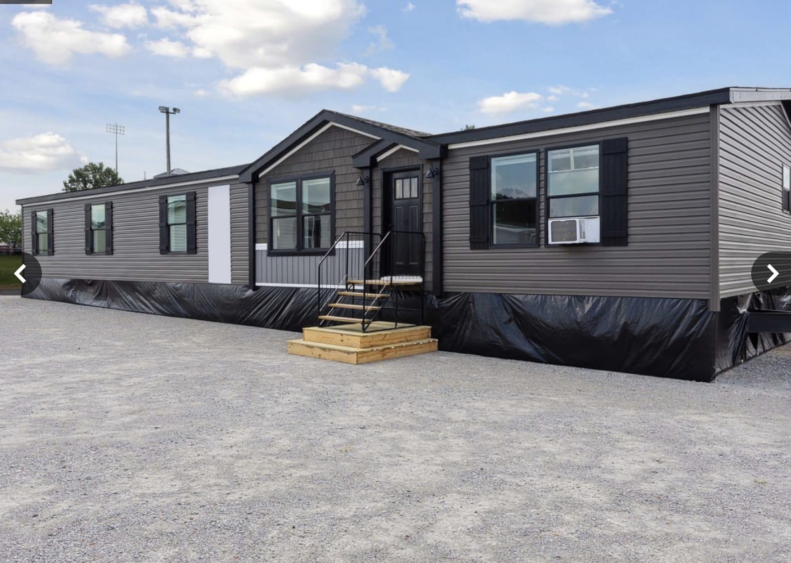 A large gray mobile home with black shutters and a small porch sits on a gravel lot under a clear blue sky. The scene is calm and tidy.