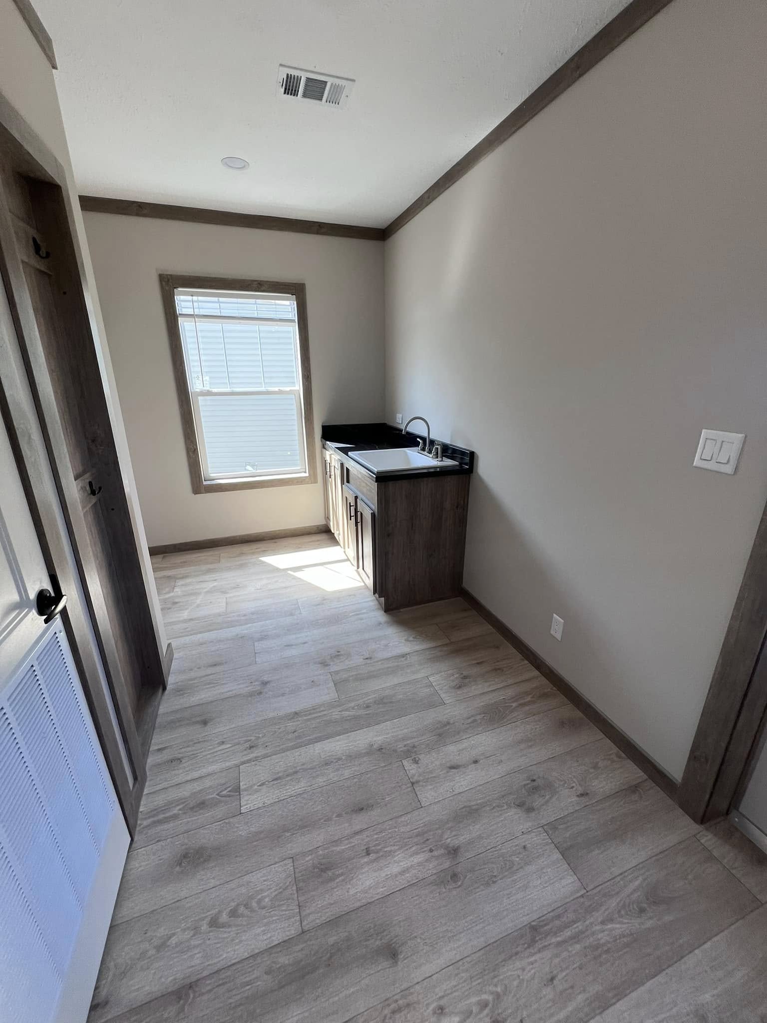 A small, narrow room with light wood flooring and beige walls. It features a single window and a compact wooden cabinet with a dark countertop and a sink. The space has a clean, minimalistic feel.