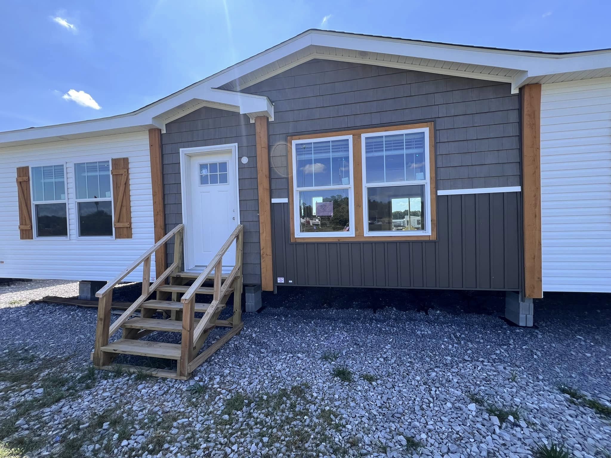Single-story modular home with a brown and white exterior, wooden stairs, and large windows. Set on gravel, it conveys a modern, welcoming feel.