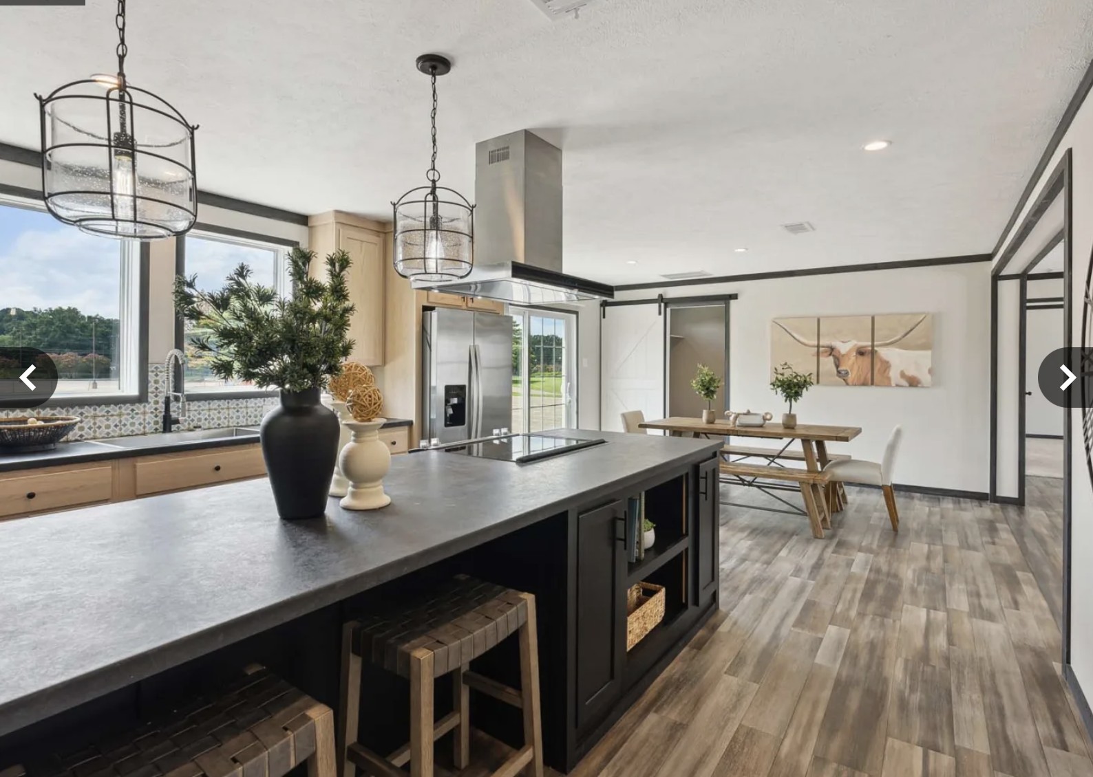 Modern kitchen with a large island featuring bar stools and a plant. Bright dining area with a wooden table and cow artwork. Neutral tones create a cozy feel.