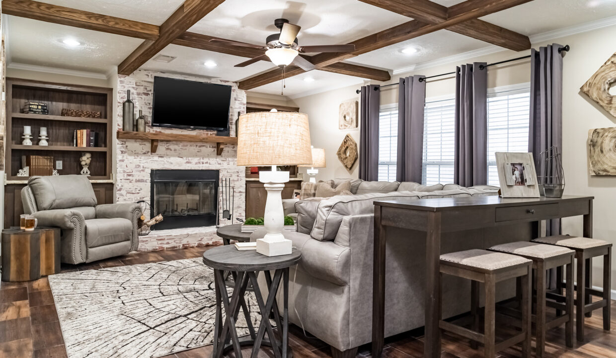 Cozy living room with wooden beam ceiling, a comfortable gray sectional, brick fireplace, bright rug, and elegant decor. Warm and inviting atmosphere.