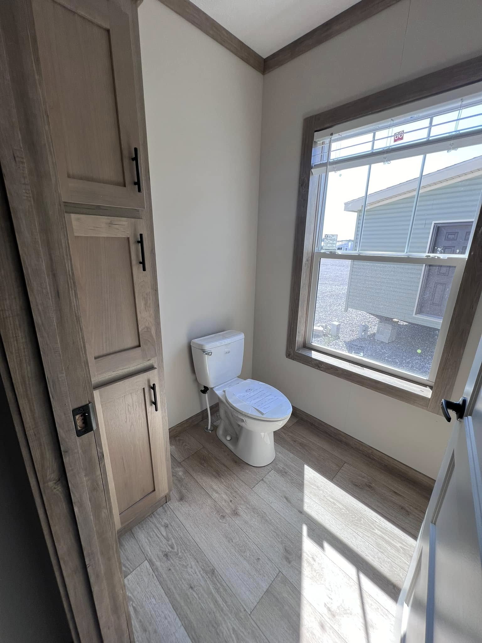 Bright bathroom with wooden cabinets and a white toilet below a sunlit window. The room has light wood flooring and a minimalist design.