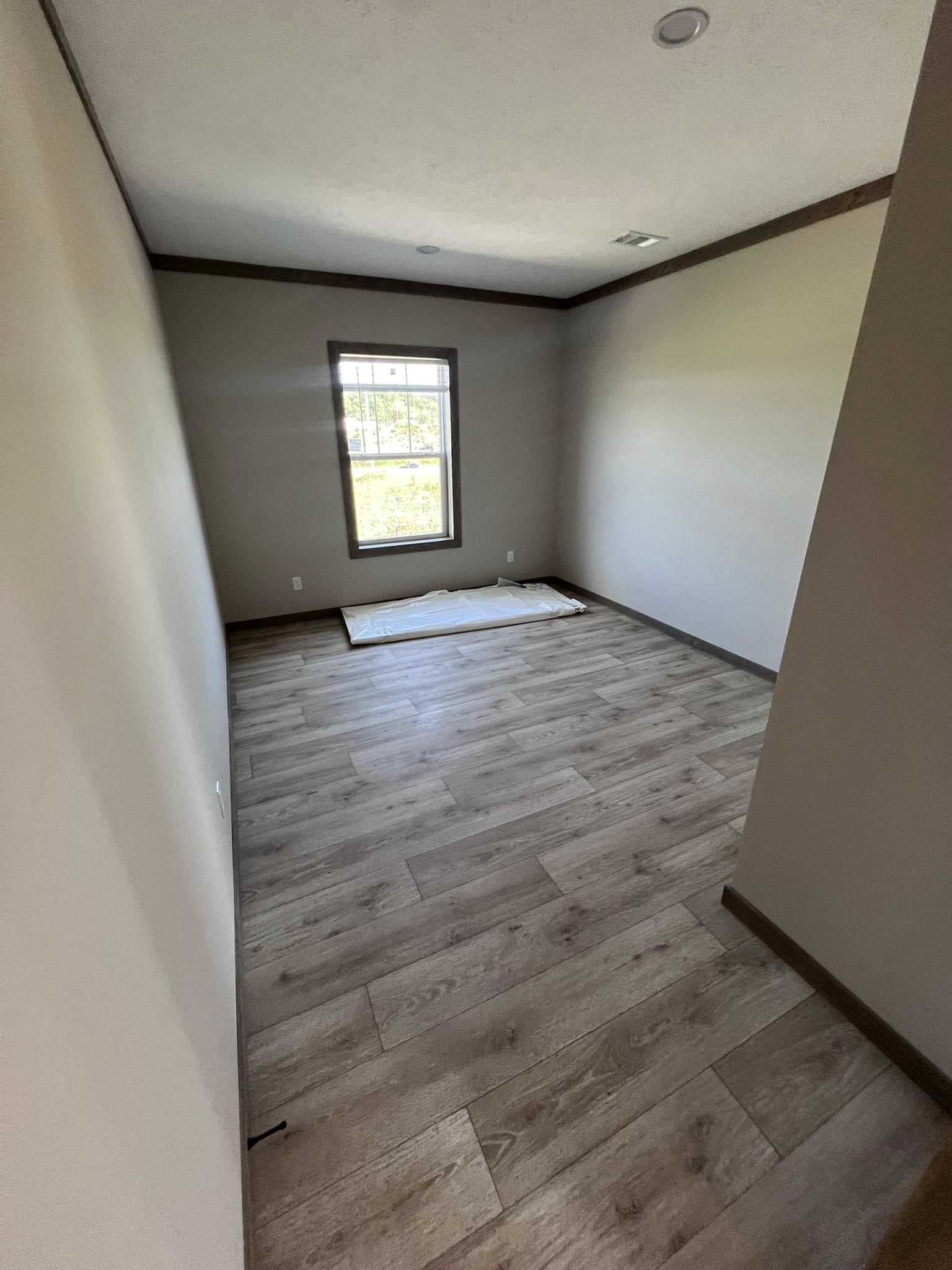 An empty room with beige walls and light wooden flooring. A single window at the far end lets in natural light, creating a calm, minimalist atmosphere.