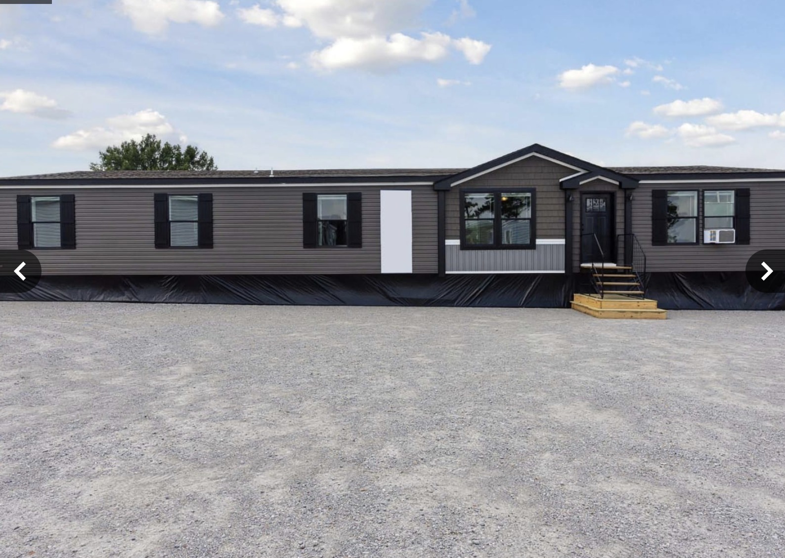 Single-story modular home with gray siding and black shutters, set against a backdrop of blue sky with clouds. A gravel driveway leads to wooden stairs.