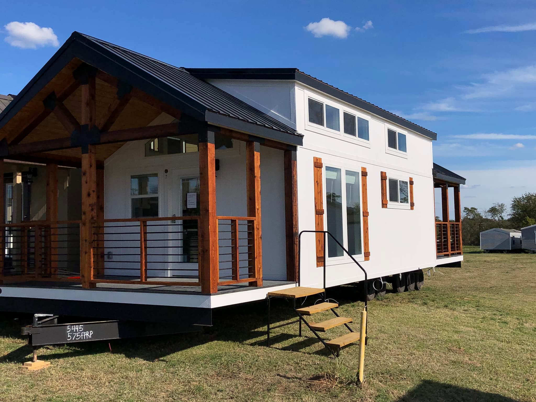 Modern tiny house with white siding and wooden accents, featuring a small porch and black metal roof. Set on grassy field under a clear blue sky.