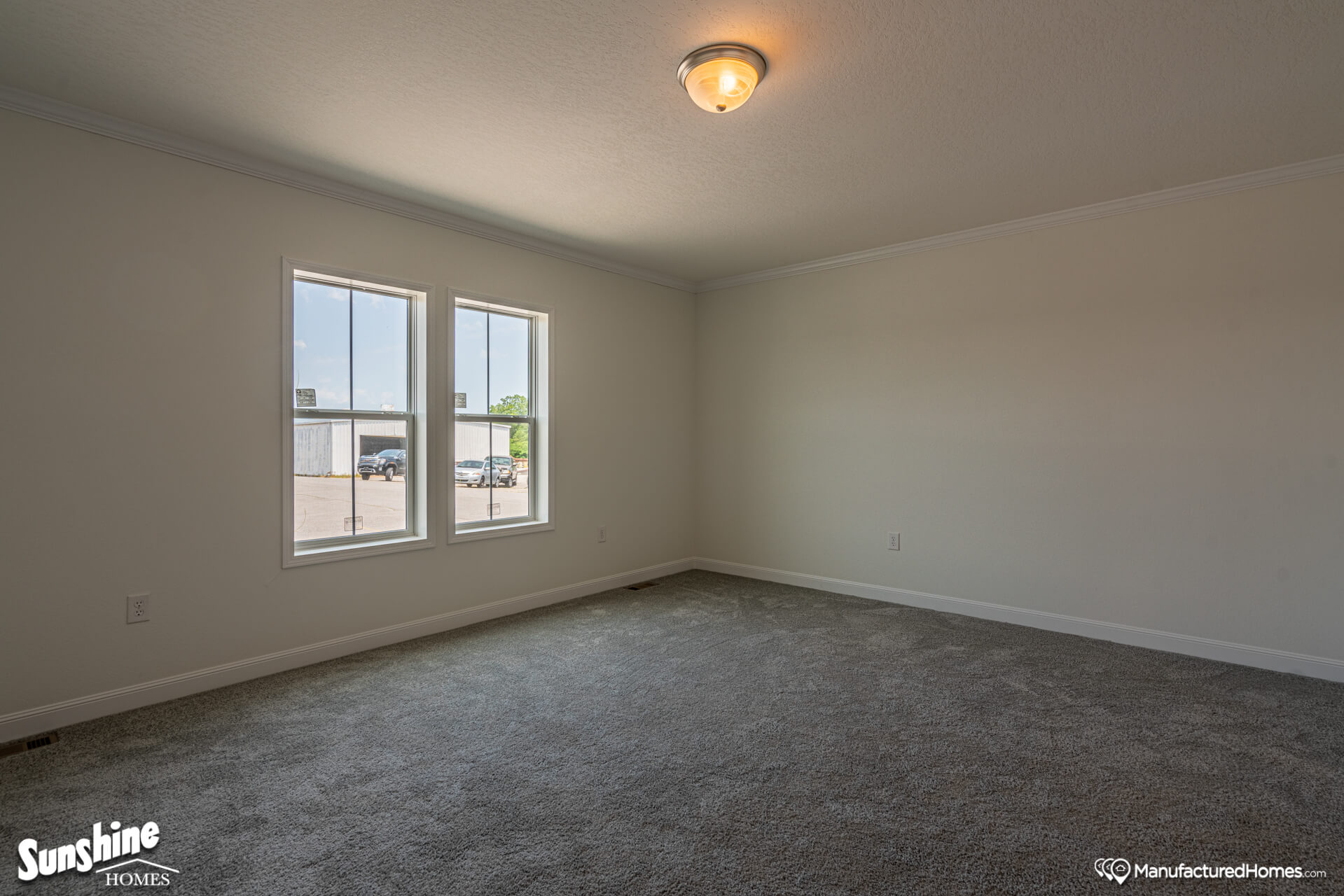 Empty room with beige walls, gray carpet, and a ceiling light. Two large windows offer natural light, showing a view of parked cars outside. Calm ambiance.