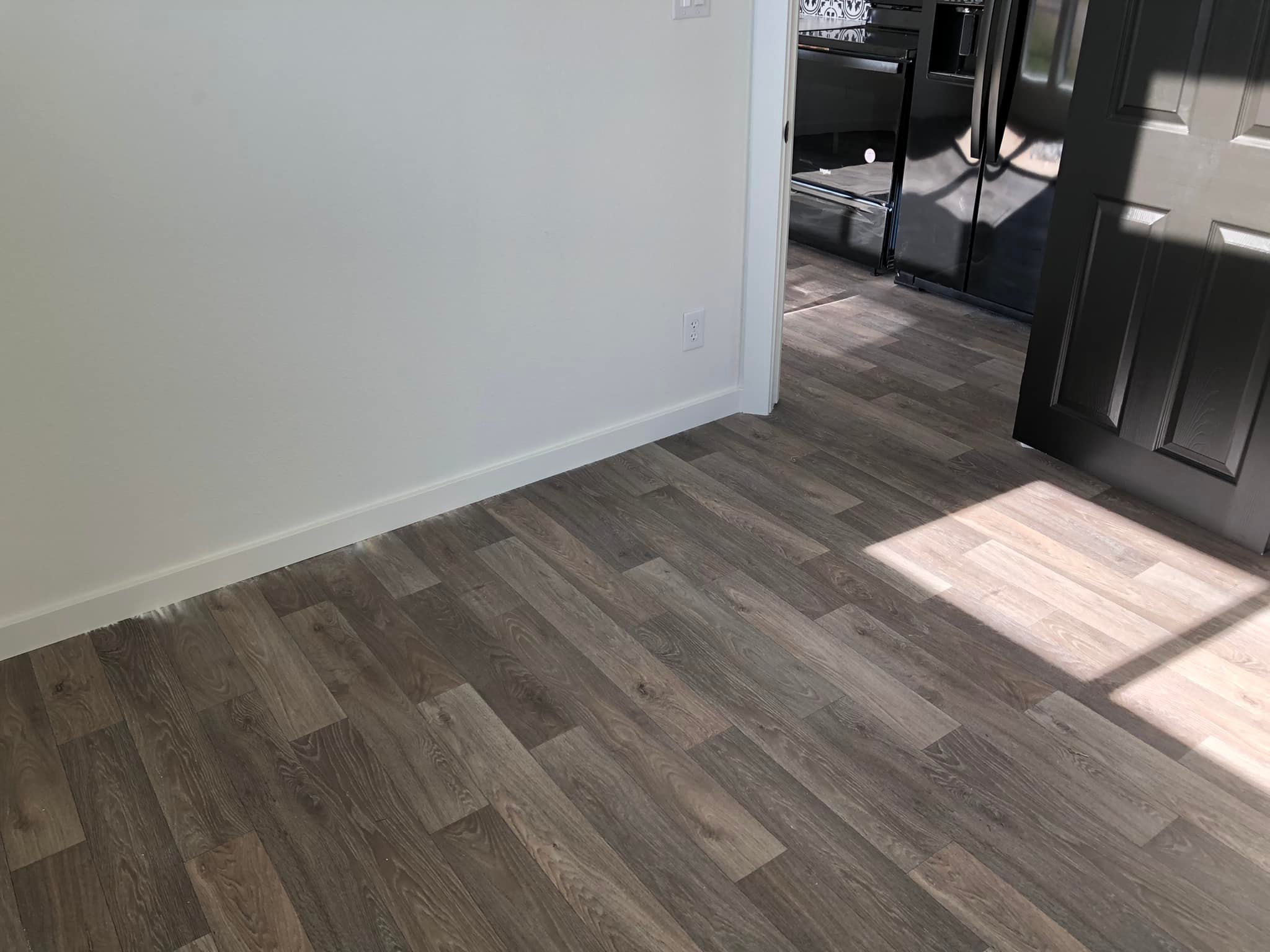 Room with light wood flooring, white walls, and a black door partially open to a kitchen. Sunlight casts shadows, creating a calm atmosphere.