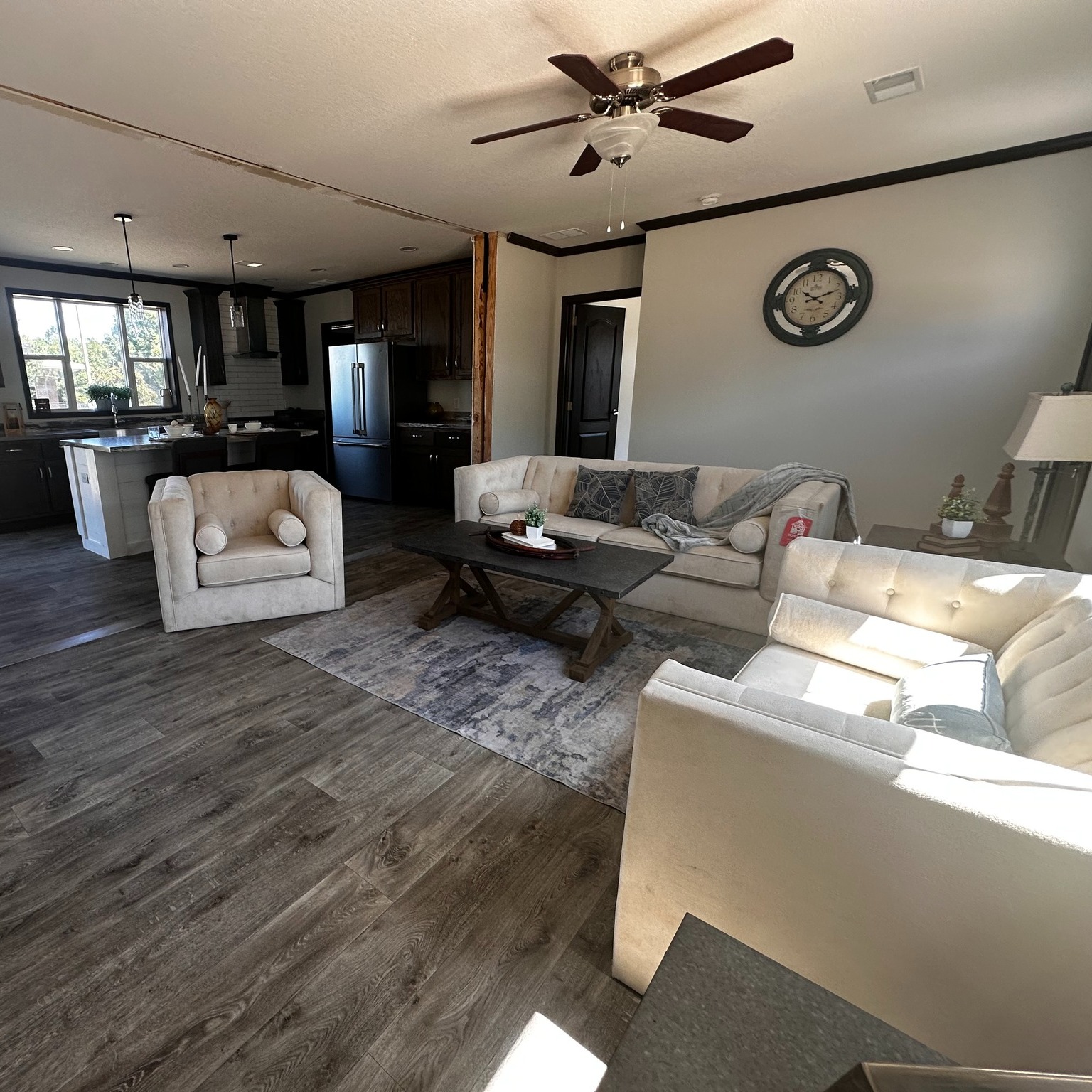 Spacious living room with beige sofas around a wooden coffee table, textured rug, and ceiling fan. Open kitchen with dark cabinets and large window. Cozy and inviting.