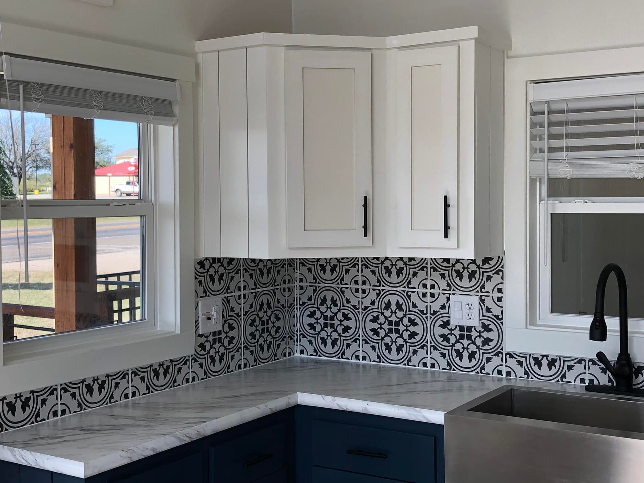 A cozy kitchen corner with white cabinets and dark handles, featuring a patterned black and white backsplash. Blue lower cabinets contrast the marbled countertop. Two windows provide natural light.
