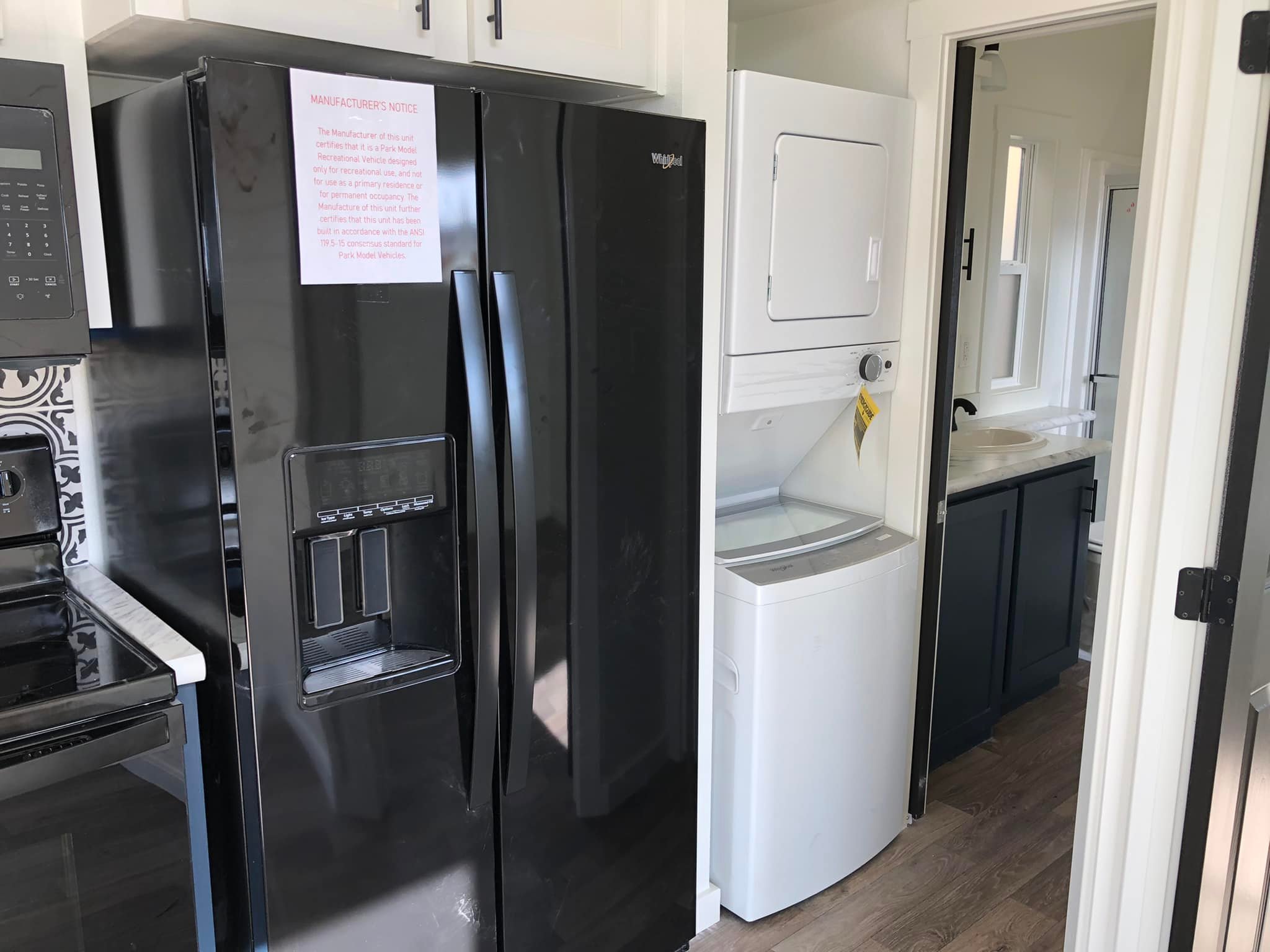 Modern kitchen with a sleek black refrigerator beside a stainless steel stove. A compact laundry area with a white stacked washer and dryer is visible, leading to a bathroom with a white sink and dark cabinetry. The space feels efficient and modern.