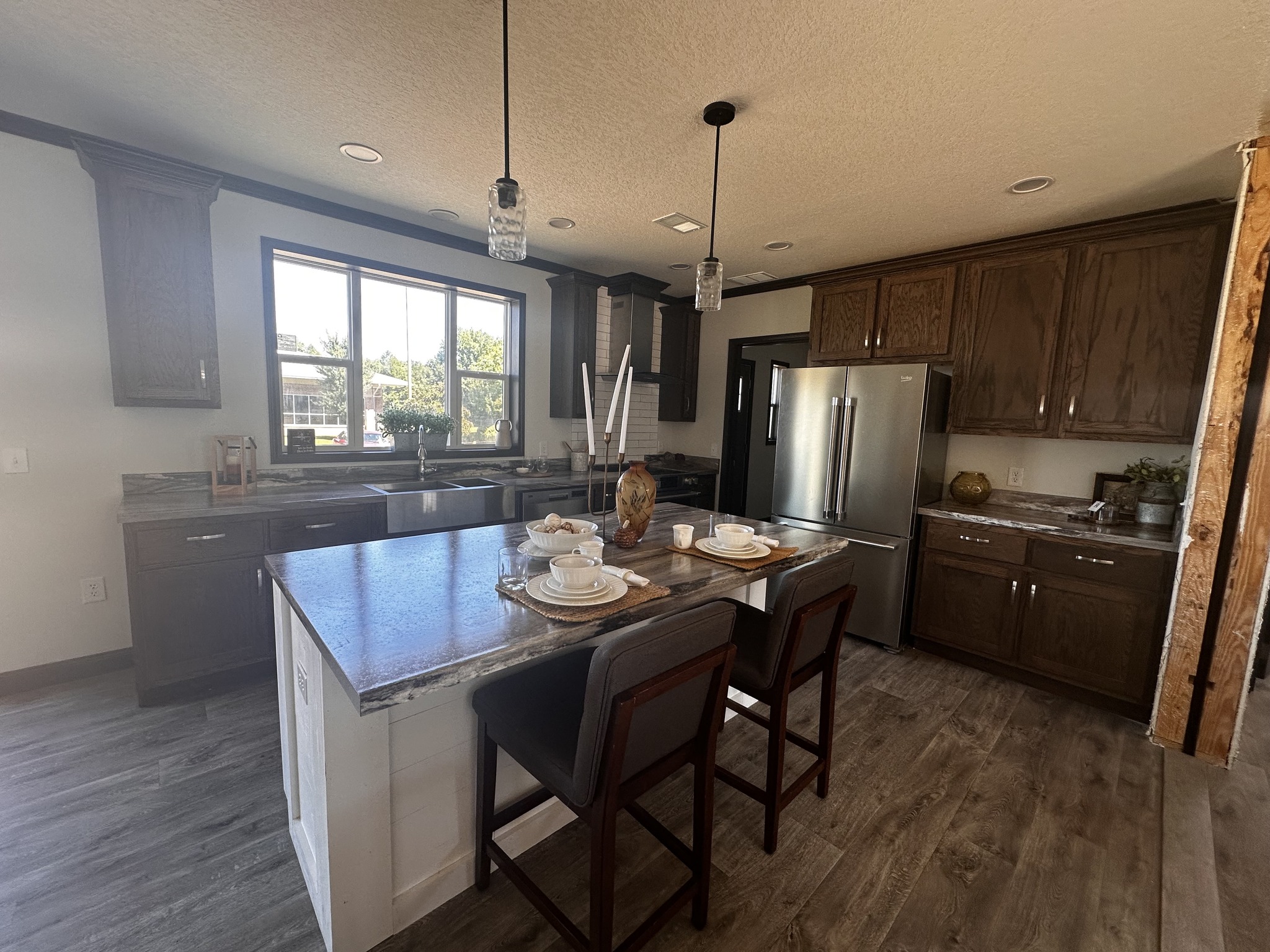 Modern kitchen with dark wood cabinets, a stainless steel refrigerator, and a central island with stools. Bright window light illuminates the space.