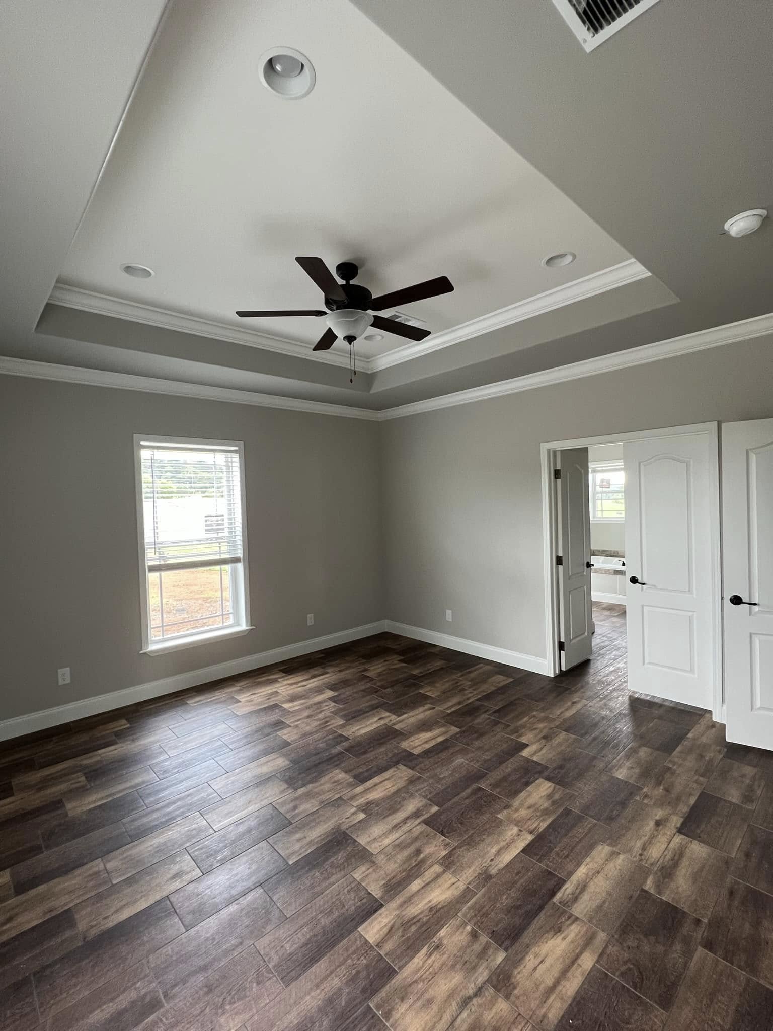 Empty room with gray walls and dark wood flooring. Features a tray ceiling with a ceiling fan, a window on the left, and a partially open white door.