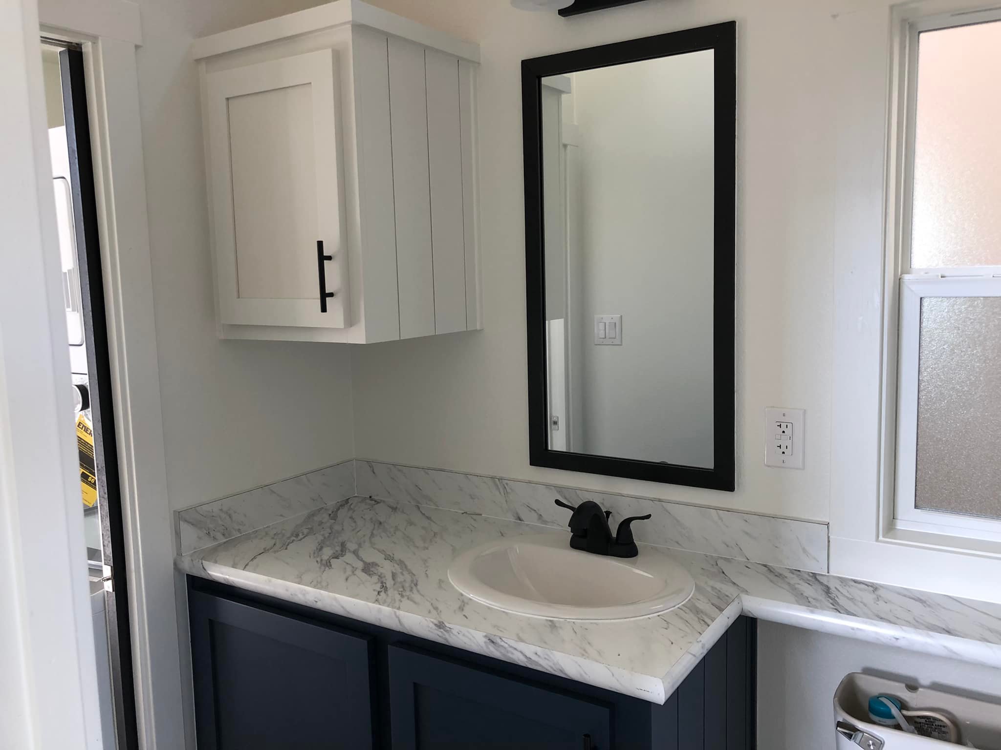 Compact bathroom corner with sleek, marble countertop, black faucet, and a rectangular mirror. White cabinet and window convey a modern, clean vibe.
