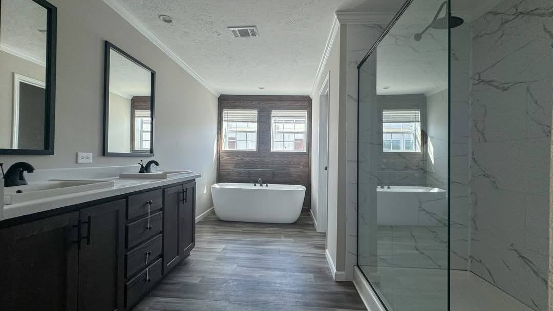 Modern bathroom with freestanding tub beneath two windows, dark wood vanity with dual sinks and mirrors, glass-enclosed shower, and wood-look flooring.