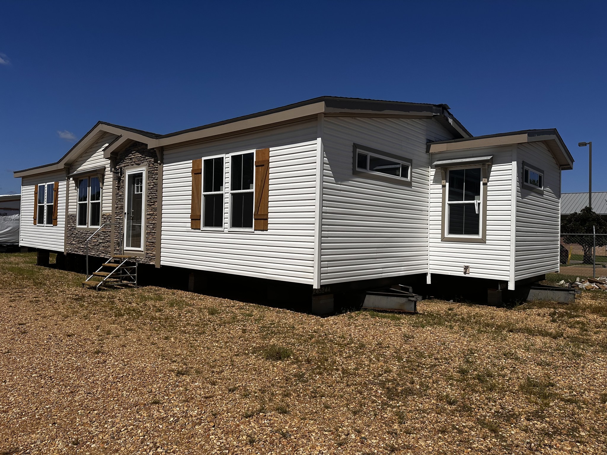 A white manufactured home is set on a gravel lot under a clear blue sky. It features wood shutters, a stone accent near the front door, and multiple windows.