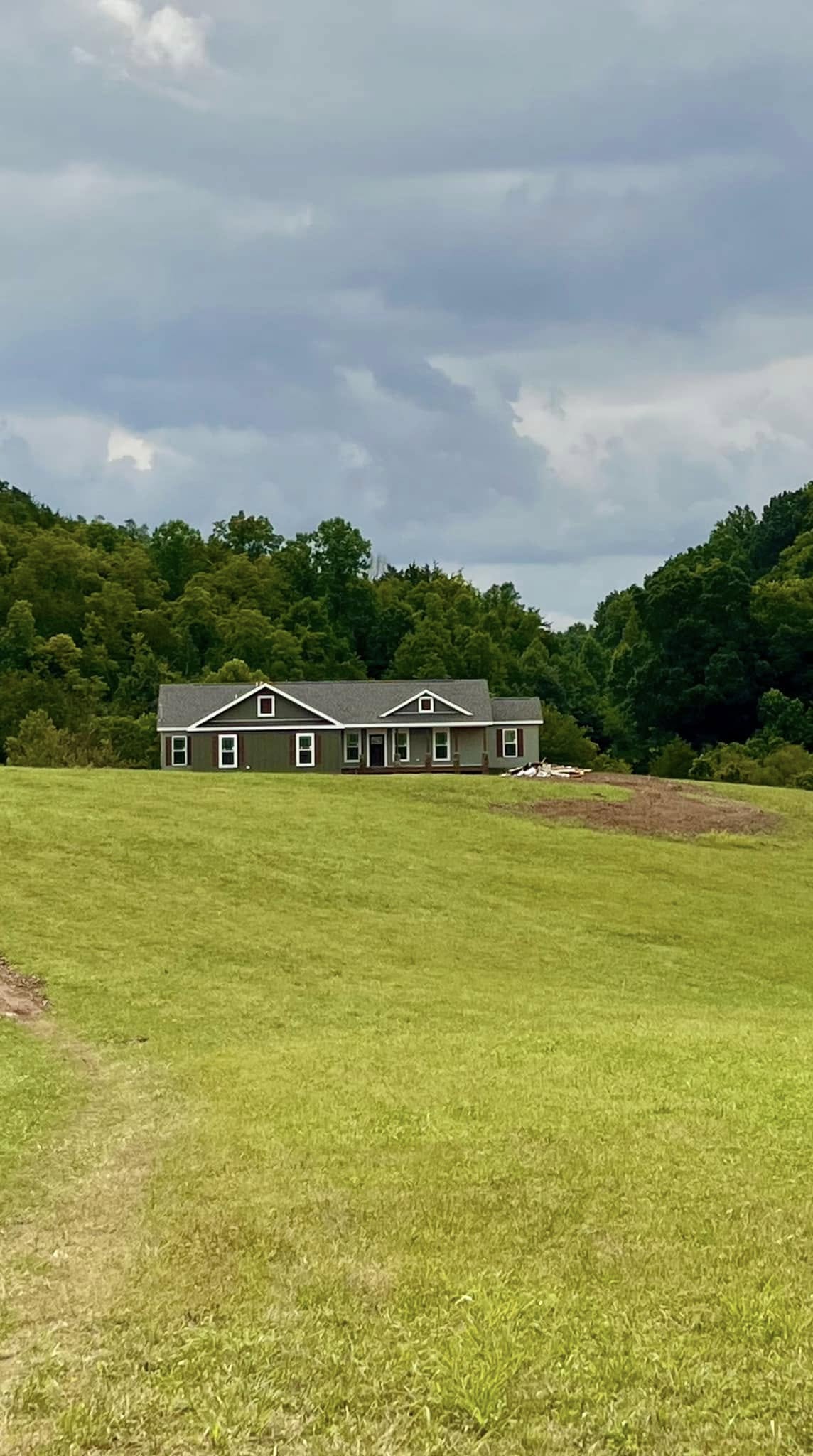 A single-story house with a gray roof sits atop a grassy hill, surrounded by lush trees under a cloudy sky, conveying a peaceful rural scene.