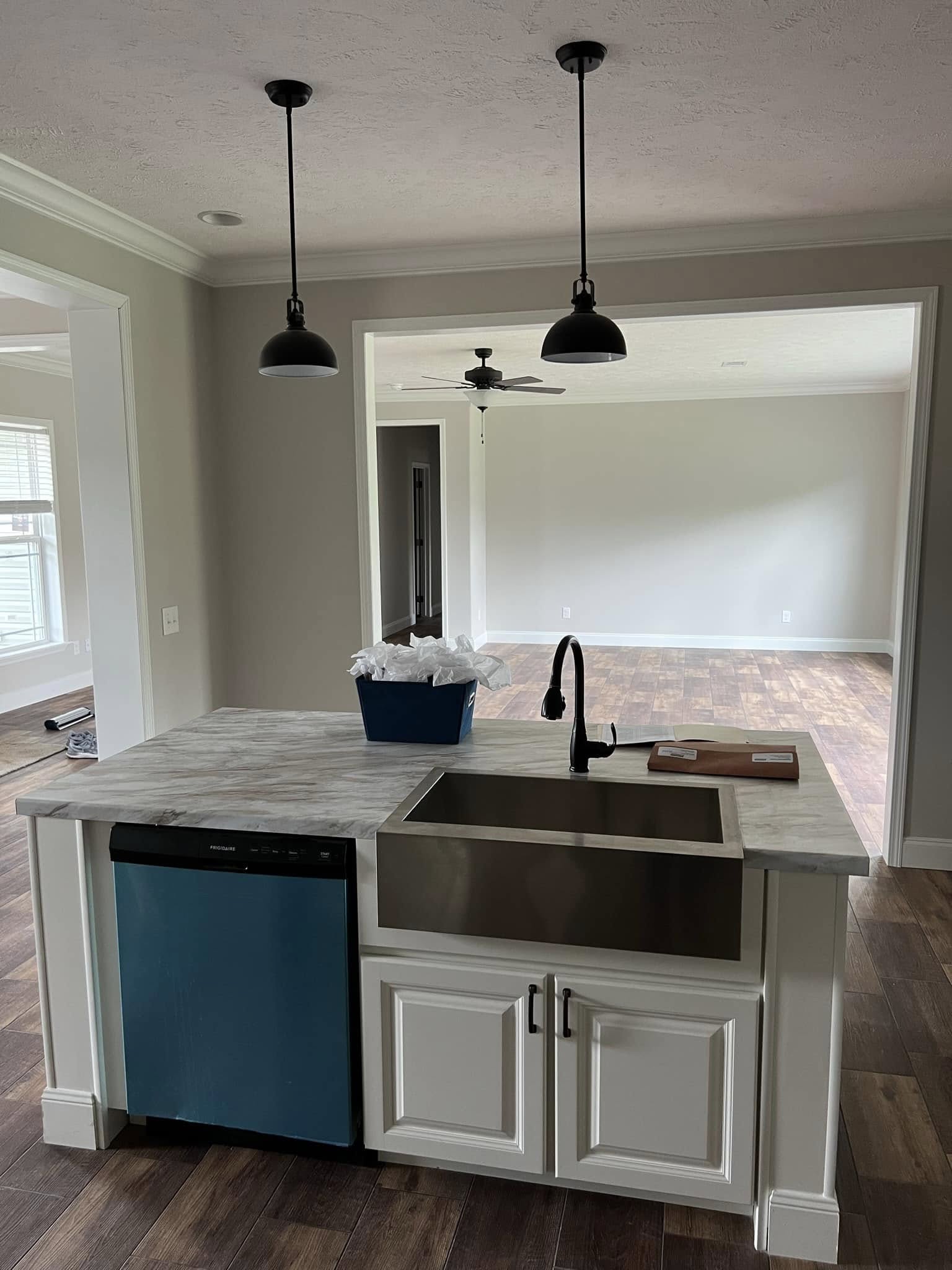 Modern kitchen with white cabinets, a farmhouse sink, and a marbled countertop. Two black pendant lights hang above. Wooden floors and neutral walls.