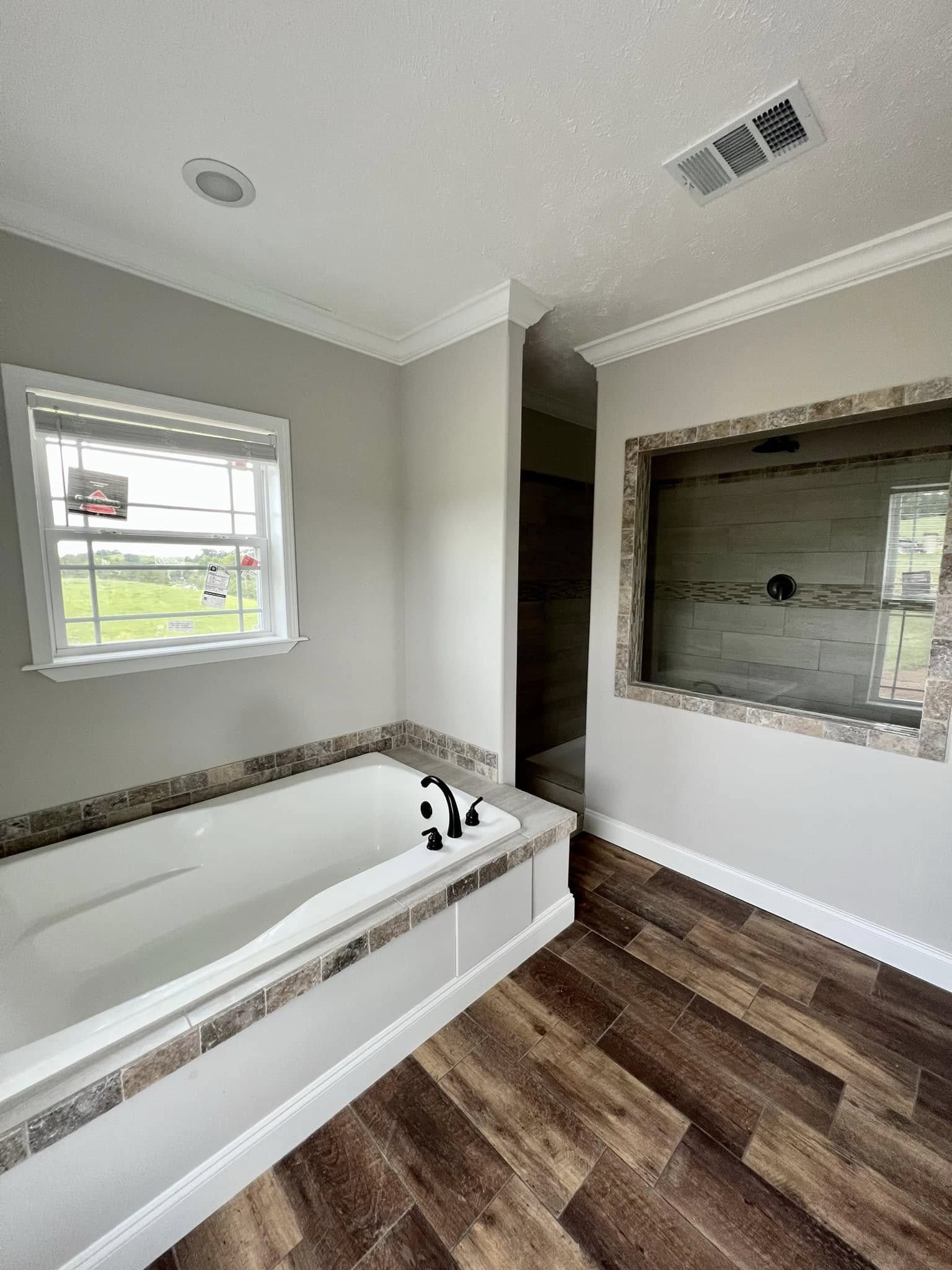 Spacious bathroom with large white tub, dark fixtures, and wood-textured tiles. Glass-enclosed shower, window with countryside view, and soft lighting.