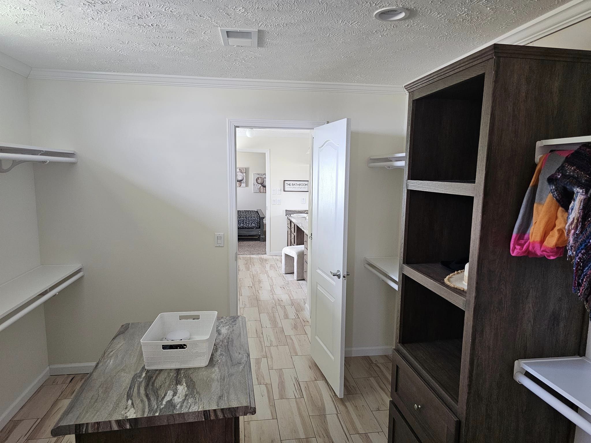 Spacious closet with open shelves and clothing rack. Marble-topped island with a basket. Open door leads to a room with a sofa. Bright, organized feel.
