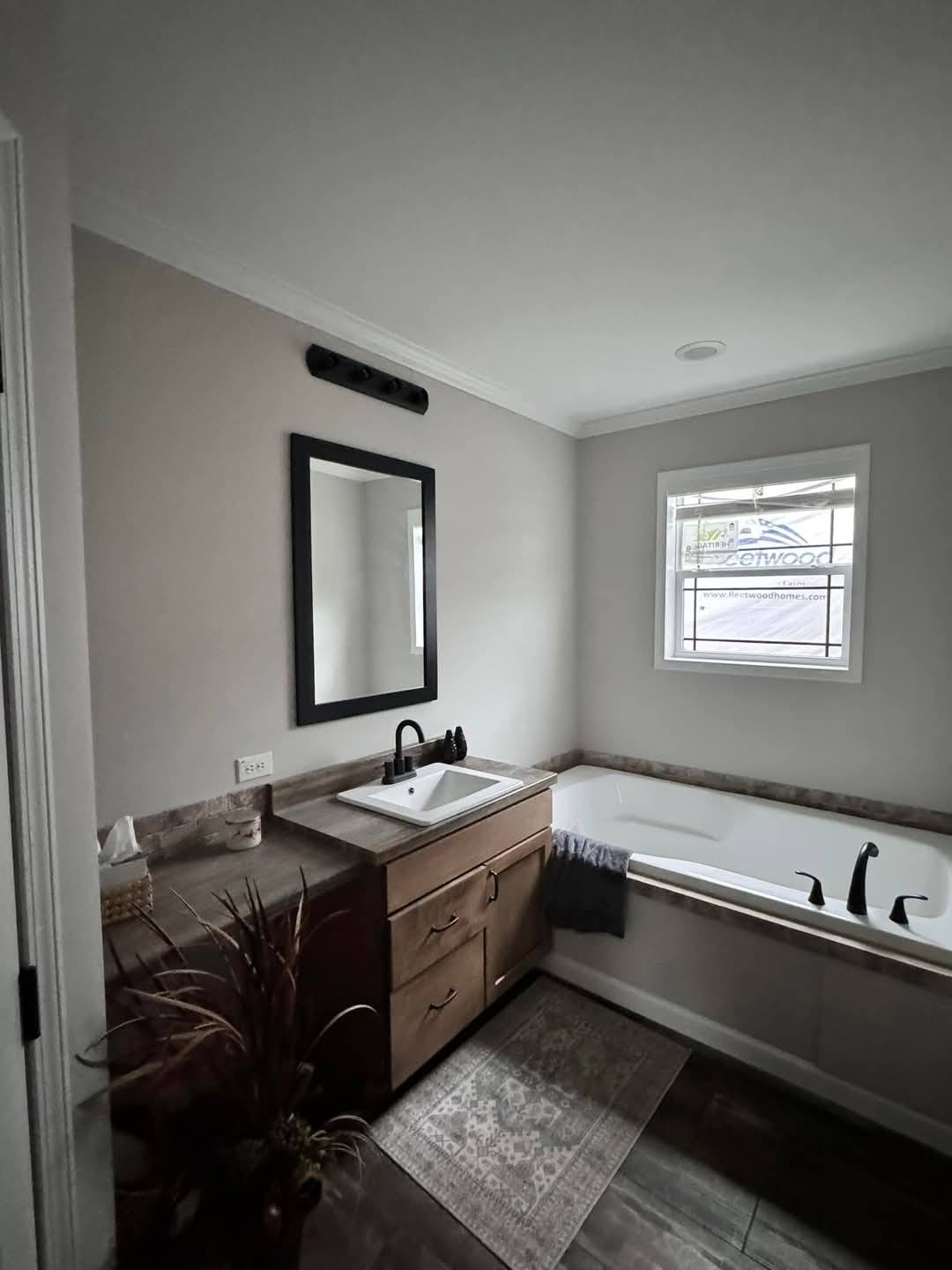 Modern bathroom with dark wood floors, a sleek wood vanity, and a black-framed mirror above a white sink. A large bathtub is near a window, creating a calm, inviting atmosphere.