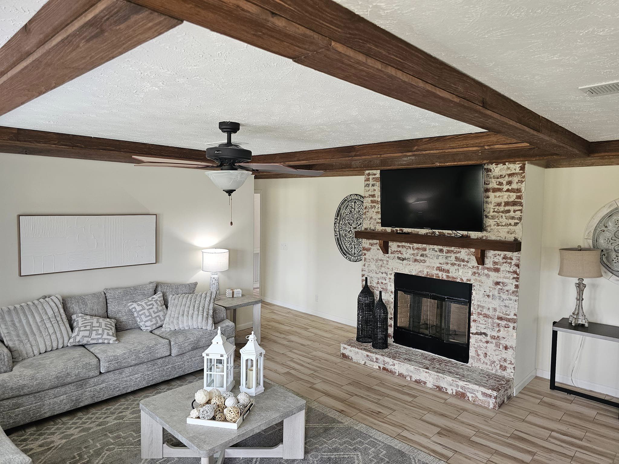 Cozy living room with rustic wood beams, a brick fireplace, and soft gray couch. Decor includes lanterns, a ceiling fan, and neutral tones.