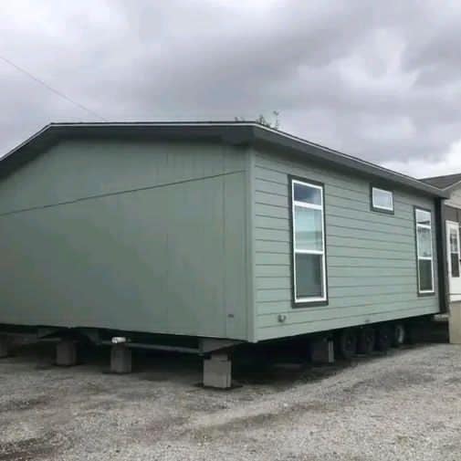 Green mobile home on concrete blocks with dark clouds overhead. The home has two windows and sits on a gravel surface, conveying a temporary setting.