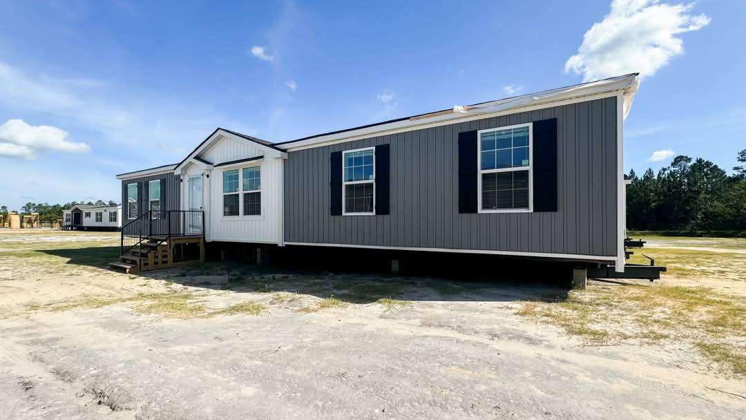 A gray and white manufactured home with black shutters sits on a sandy lot under a clear blue sky. Steps lead to the main door. Trees are visible in the distance.