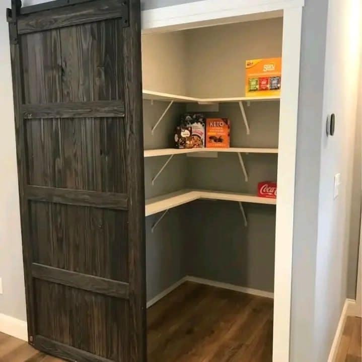 Rustic wooden sliding door partially open, revealing a small, organized pantry with two white shelves stocked with cereal boxes, snacks, and a soda pack.