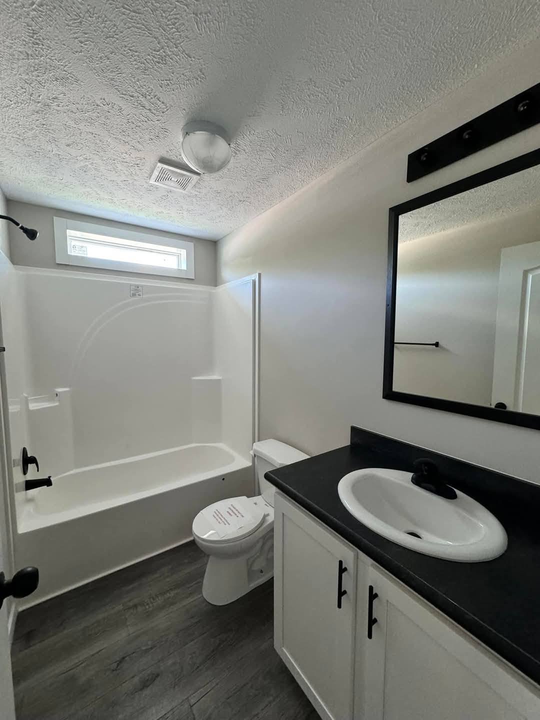 A modern bathroom with a white bathtub and shower, a black-countered sink, and a framed mirror. The space is neat and bright with a window above.