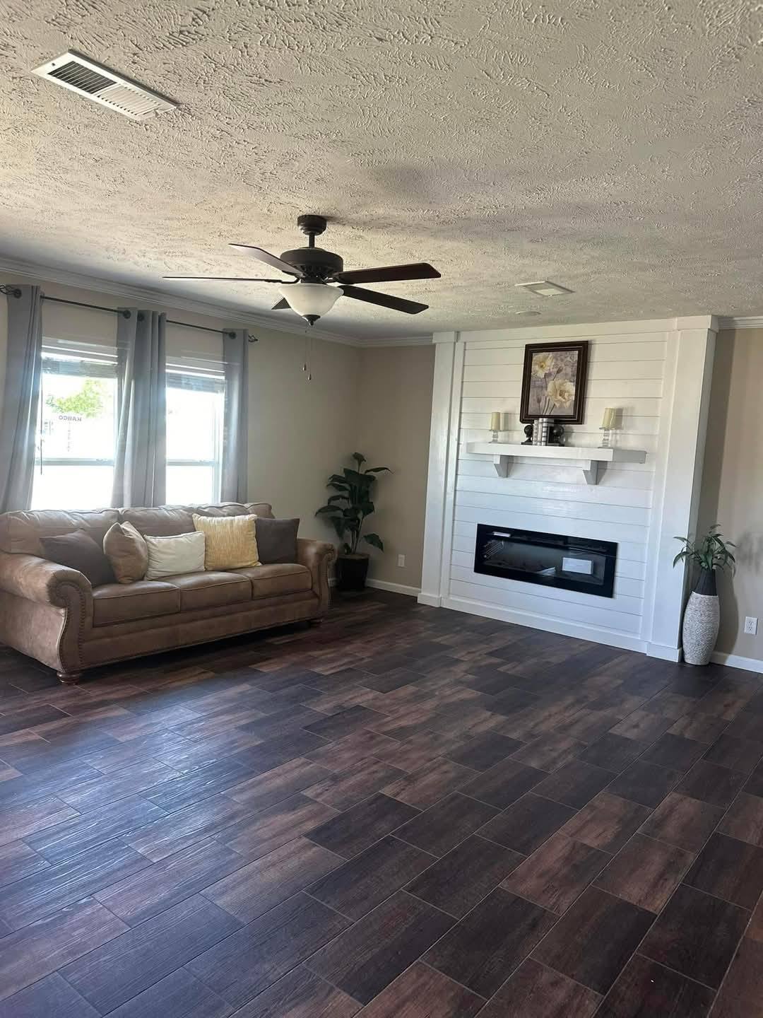A cozy living room with a brown couch adorned with pillows, wood-look flooring, ceiling fan, and a white shiplap fireplace with decor, conveying warmth.