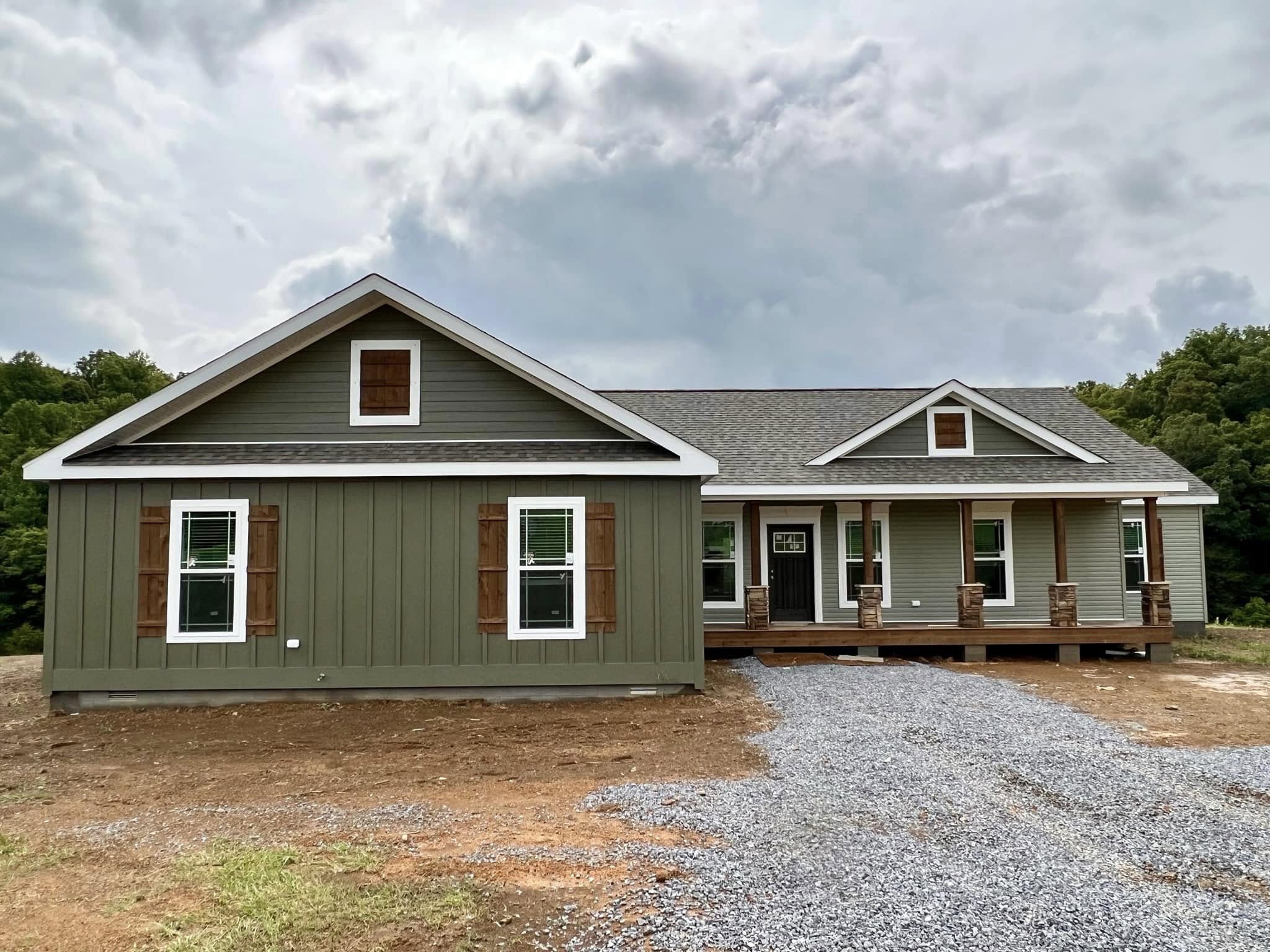 A single-story green house with brown shutters and a gravel driveway stands against a cloudy sky. The porch has wooden pillars, conveying a rustic feel.