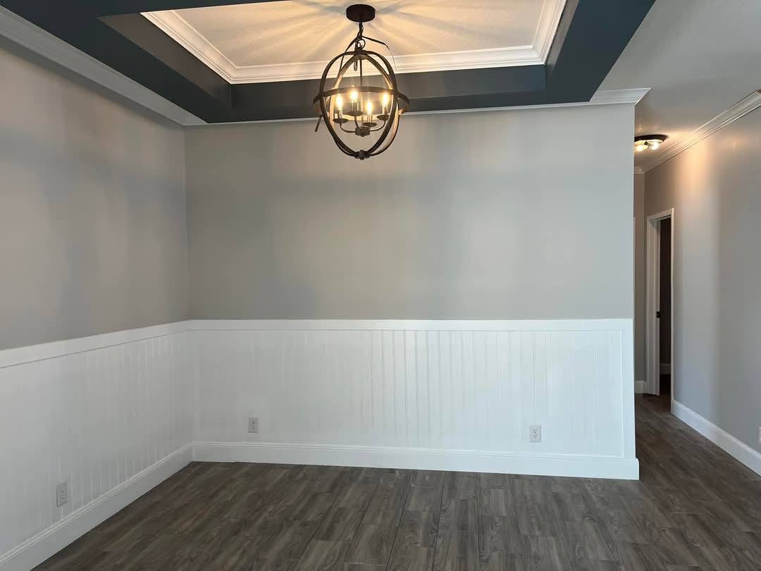 Empty room with light gray walls and white wainscoting. A spherical chandelier hangs from a coffered ceiling. Dark wood flooring adds warmth.