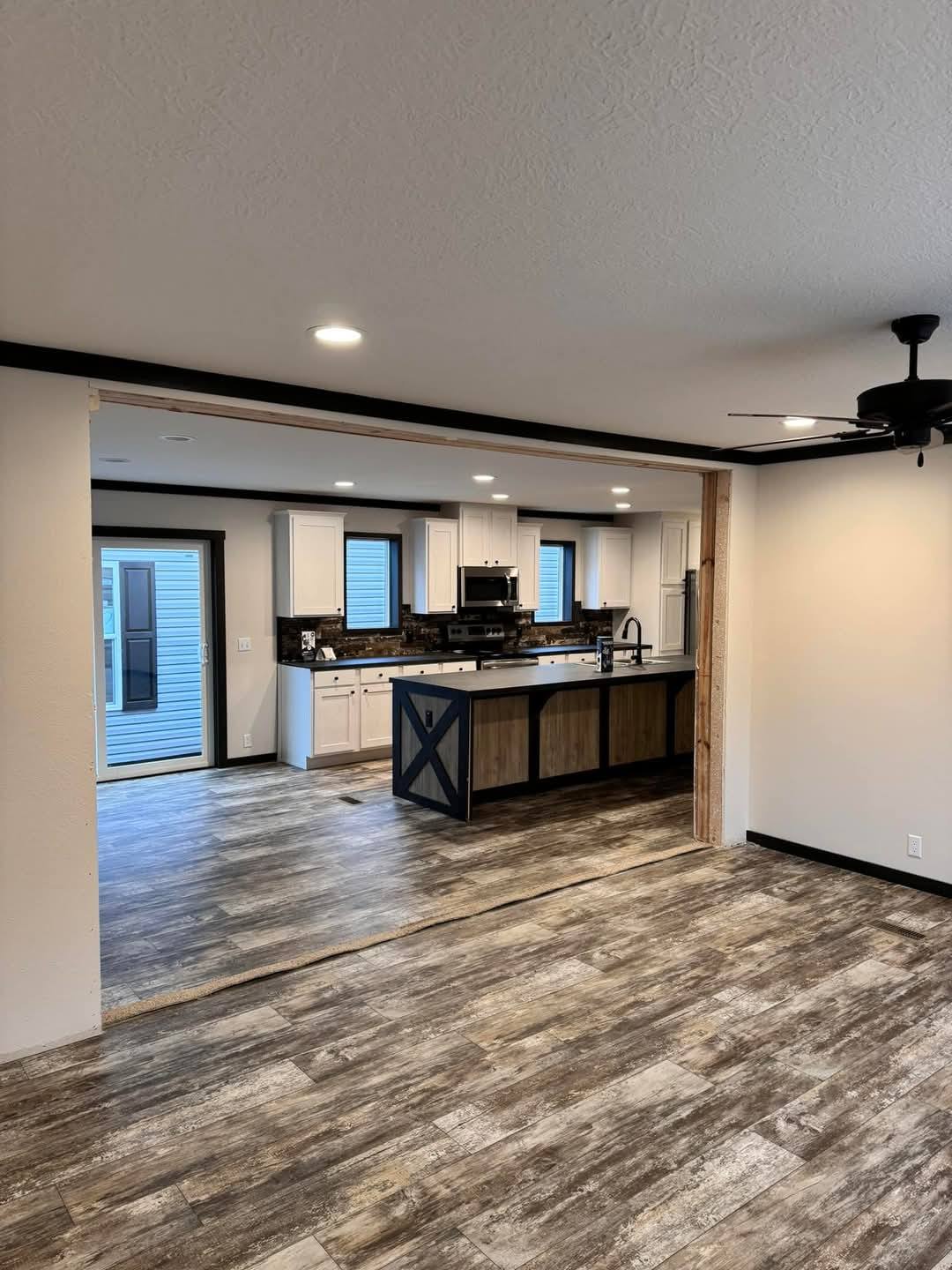 Modern open-concept kitchen and living area with rustic wood flooring, white cabinets, black countertop, and ceiling fan. Bright, spacious, inviting.