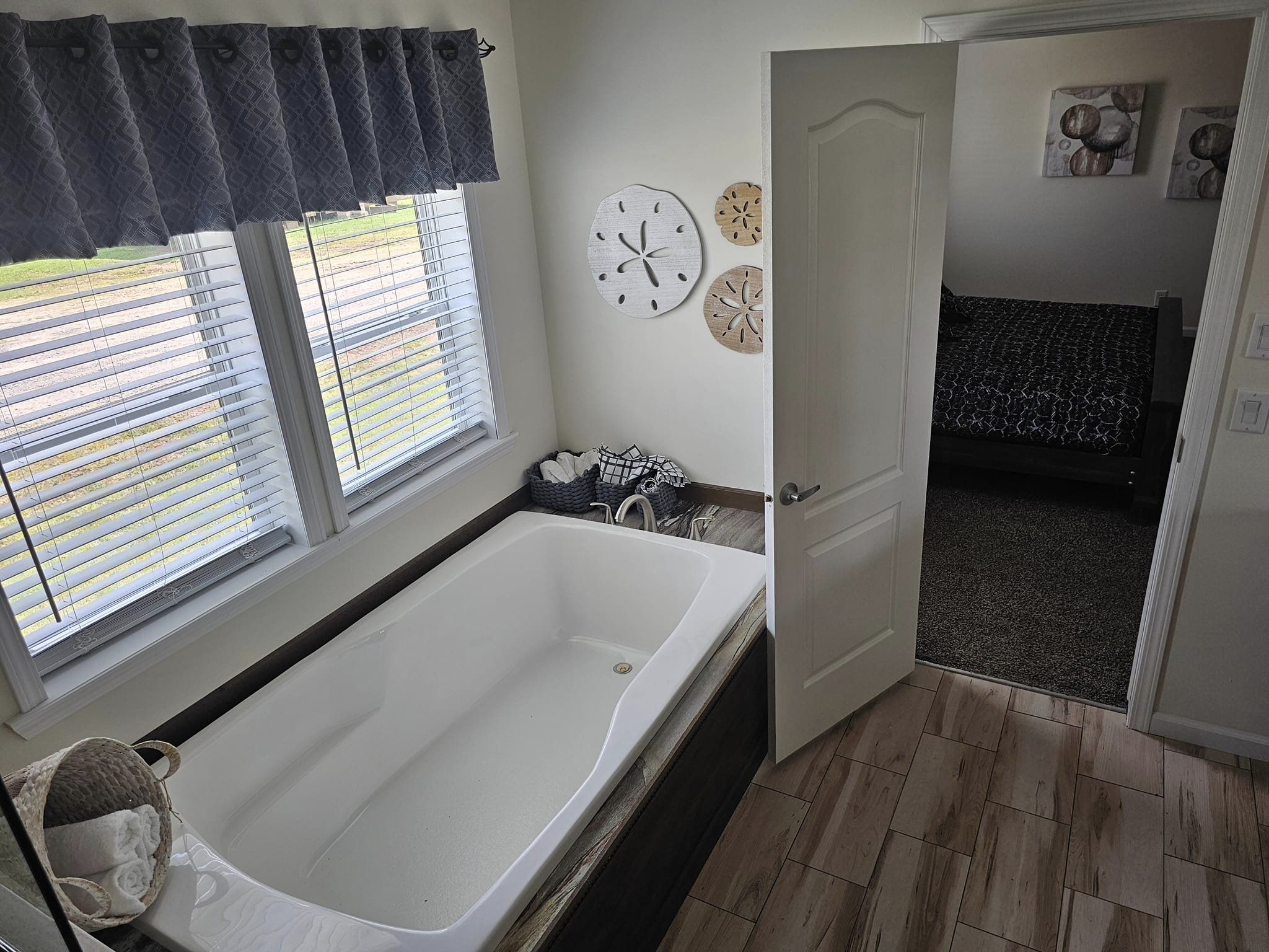 Modern bathroom with a white bathtub under a window with gray curtains. Open door leads to a bedroom. Walls feature circular decor, tone is serene.