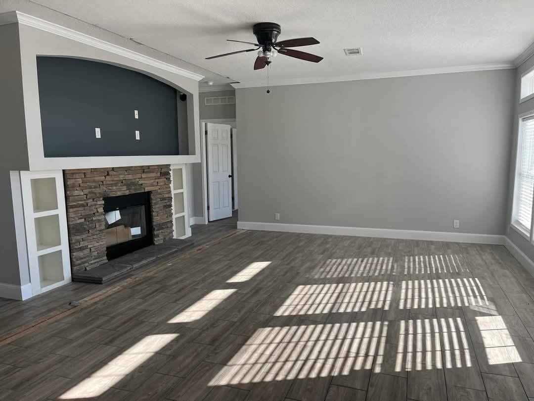 Spacious living room with gray walls, wooden floor, and a stone fireplace. Light filters through large windows, casting striped shadows.