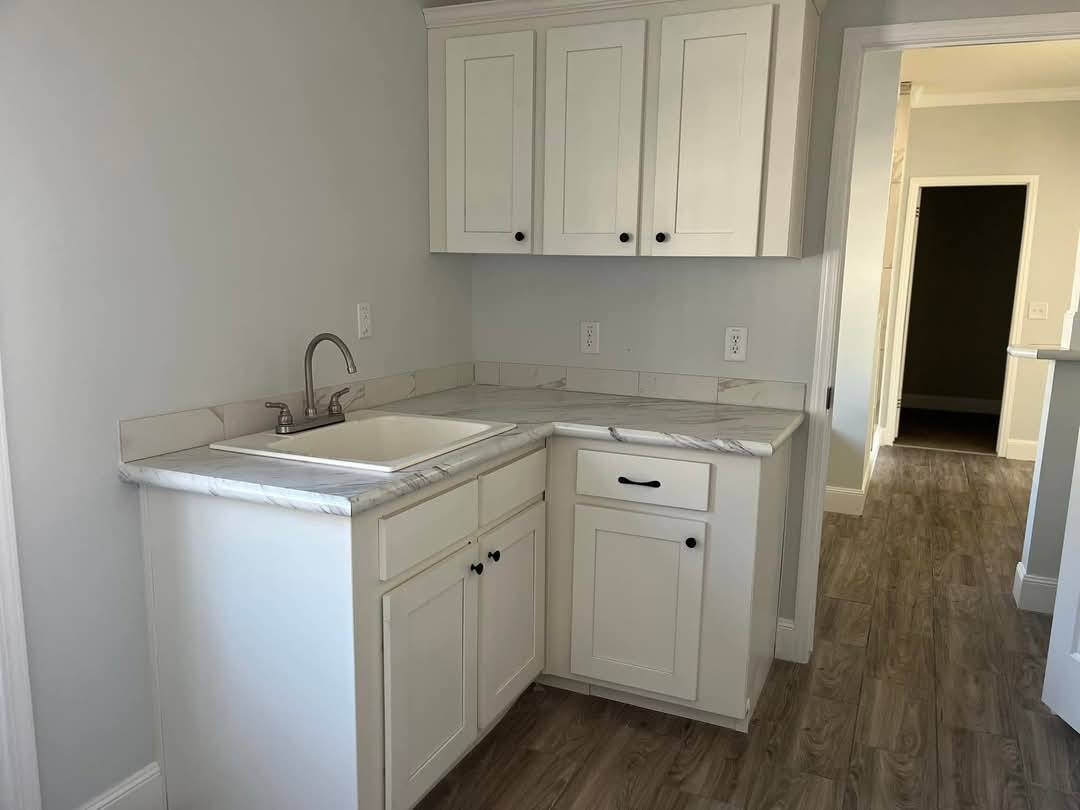 Small kitchen area with white cabinets, marble countertops, and a stainless steel faucet over a white sink. It has wood flooring and light gray walls.