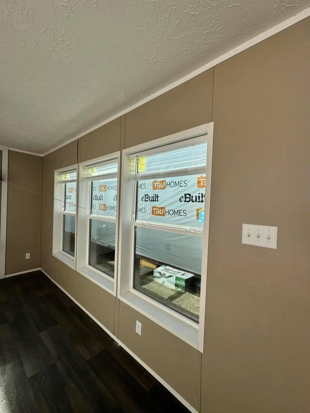 Interior of a room with three large windows showing "TRU Homes" branding outside. Beige walls, dark wood flooring, and a light switch on the wall.