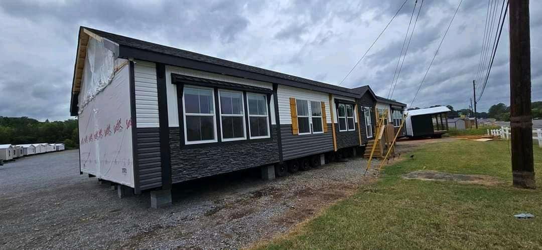 A long, dark gray manufactured house with multiple windows and wooden shutters is set on a grassy area under a cloudy sky, conveying a calm atmosphere.