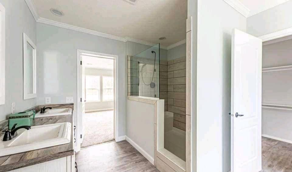 Spacious bathroom with dual sinks, marble counters, and a walk-in shower. Light gray walls, wood floors, and open doors leading to a bright room.