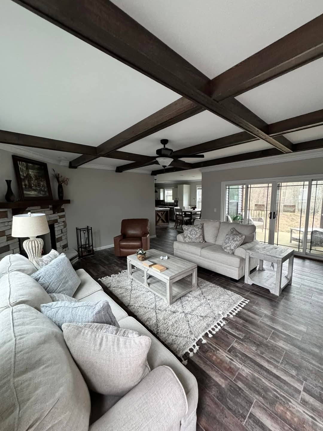 Spacious living room with beige sofas, a patterned rug, and wooden ceiling beams. Sunlit sliding glass doors lead to a garden, creating a cozy, inviting ambiance.